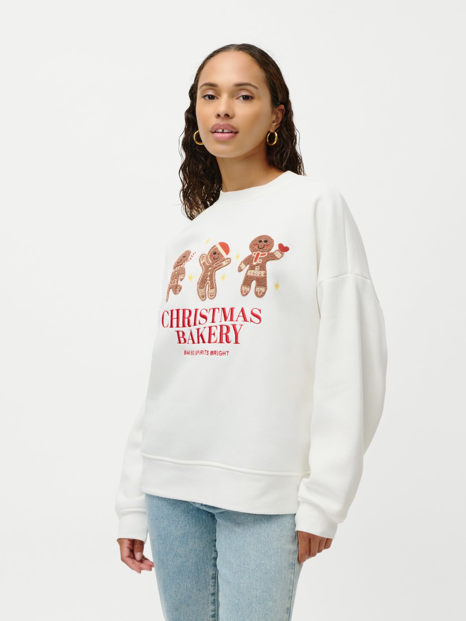 Eine Frau mit Creolen und dem weißen „Daphne“-Pullover von LeGer by Lena Gercke, der mit Lebkuchenfiguren, dem roten Schriftzug „CHRISTMAS BAKERY“, langen Ärmeln und einer lockeren Passform versehen ist, steht vor einem schlichten weißen Hintergrund.