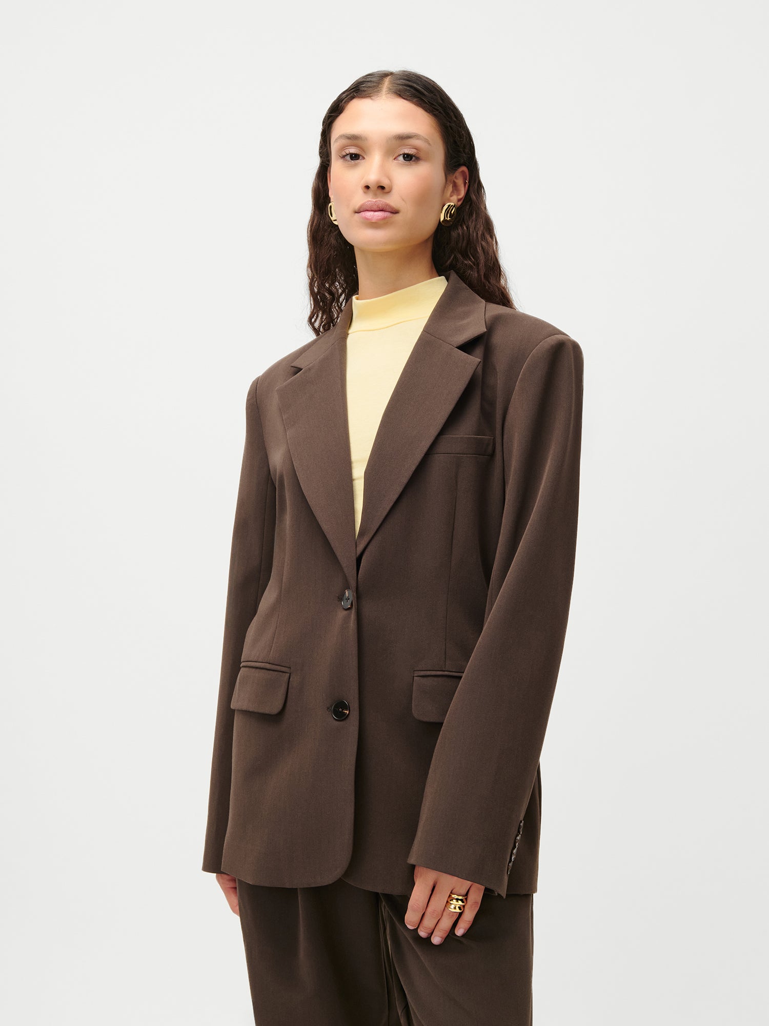 Eine Person mit gewellten dunklen Haaren trägt den braunen LeGer x Purelei - Athina Blazer von LeGer by Lena Gercke in Größe 36 über einem hellgelben Rollkragenpullover, der mit goldenen Ohrringen und Ringen verziert ist, vor einem schlichten weißen Hintergrund.