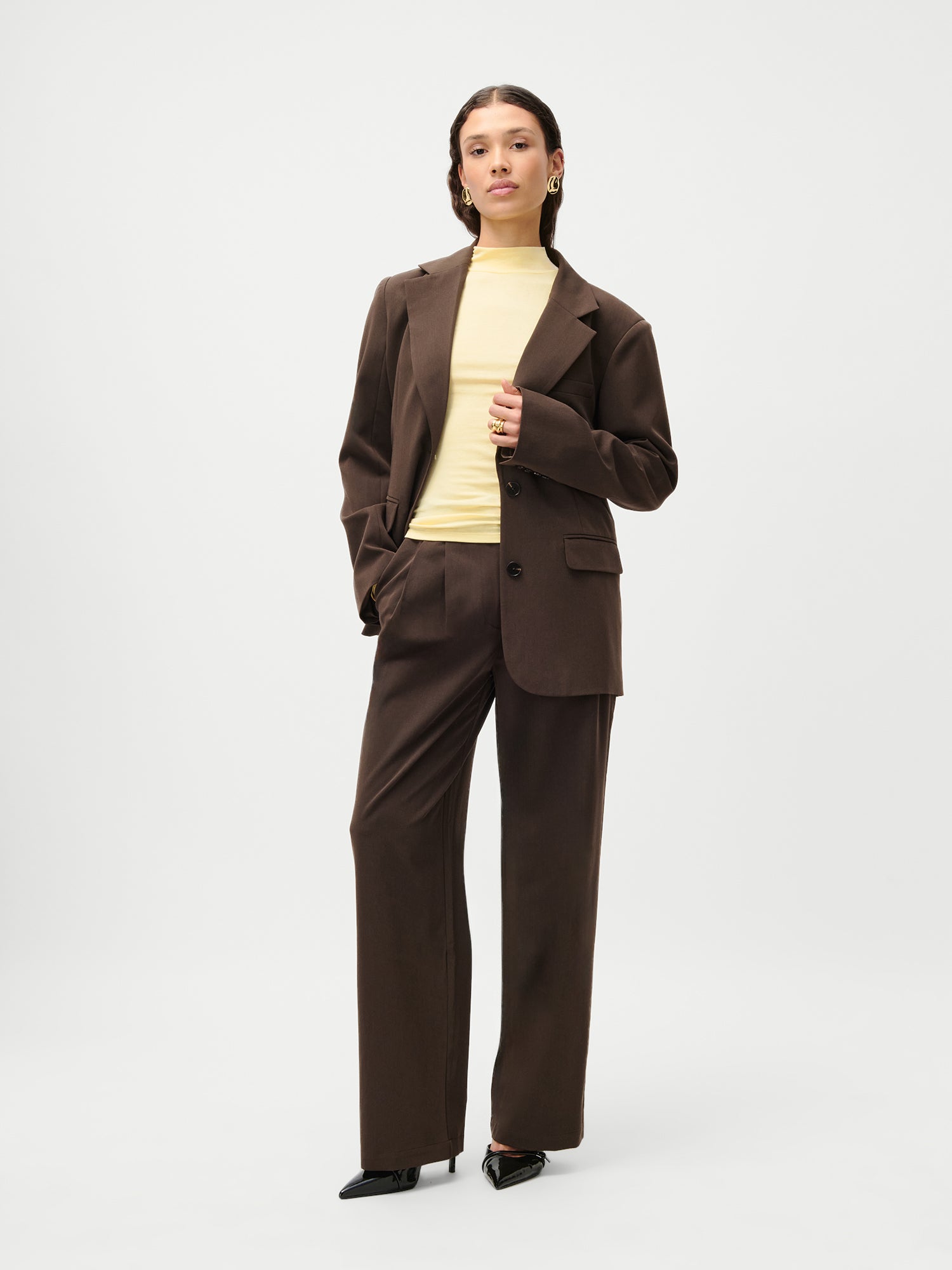 Eine Person steht vor einem weißen Hintergrund und trägt den LeGer x Purelei - Athina Blazer von LeGer by Lena Gercke in Größe 36 (normale Länge, lange Ärmel), dazu eine Hose mit weitem Bein, einen hellgelben Rollkragenpullover und schwarze spitze Absätze.