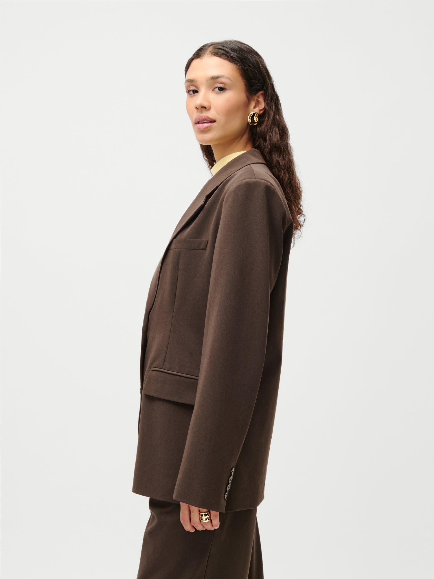 Eine Frau mit langen, gewellten Haaren trägt den LeGer x Purelei - Athina Blazer von LeGer by Lena Gercke in Größe 36 und goldene Ohrringe. Sie steht im Profil vor einem weißen Hintergrund. Der Blazer hat eine normale Länge. Sie blickt ruhig in die Kamera, die Hände an den Seiten.