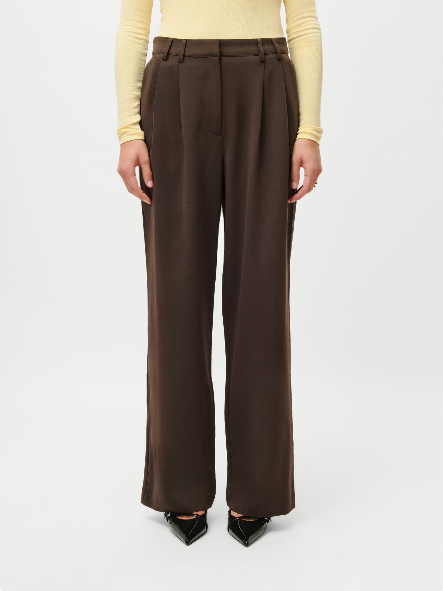 Jemand trägt die LeGer x Purelei - Athina Pants by LeGer by Lena Gercke in Braun (Größe 36), gepaart mit einem hellgelben, langärmeligen Oberteil und schwarzen, spitzen Schuhen, vor einem schlichten weißen Hintergrund.