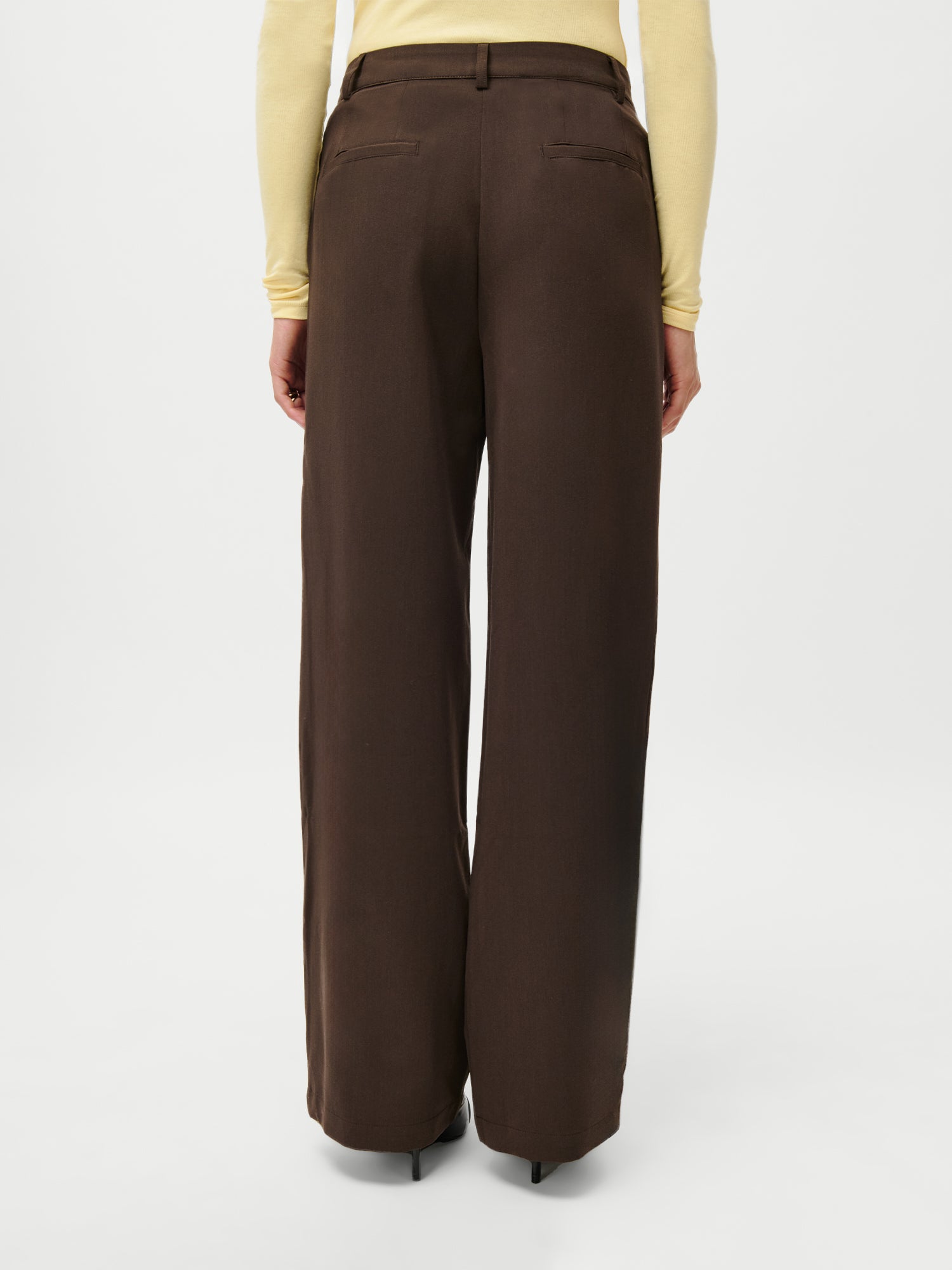 Ein Model ist von hinten vor weißem Hintergrund zu sehen. Es trägt die LeGer x Purelei Athina Pants von LeGer by Lena Gercke, eine braune Hose mit hoher Taille und weitem Bein, sowie ein hellgelbes, langärmeliges Oberteil in Größe 36.