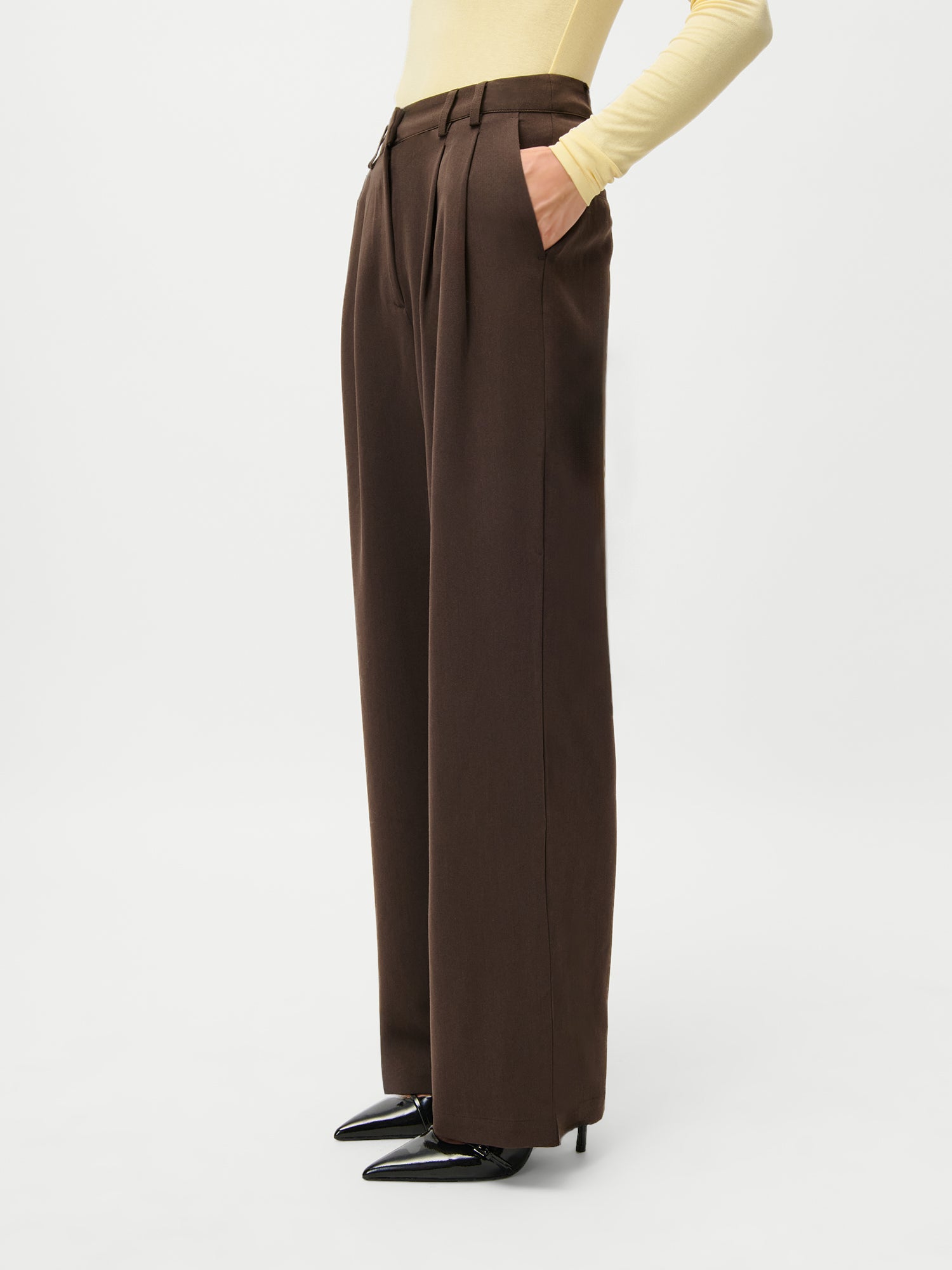 Ein Model in LeGer x Purelei - Athina Pants by LeGer by Lena Gercke, gepaart mit einem hellgelben, langärmeligen Oberteil und schwarzen, spitzen Absätzen, steht mit den Händen in den Taschen und bietet Größe 36 Style-Inspiration für Liebhaber langer Kleider vor einem weißen Hintergrund.