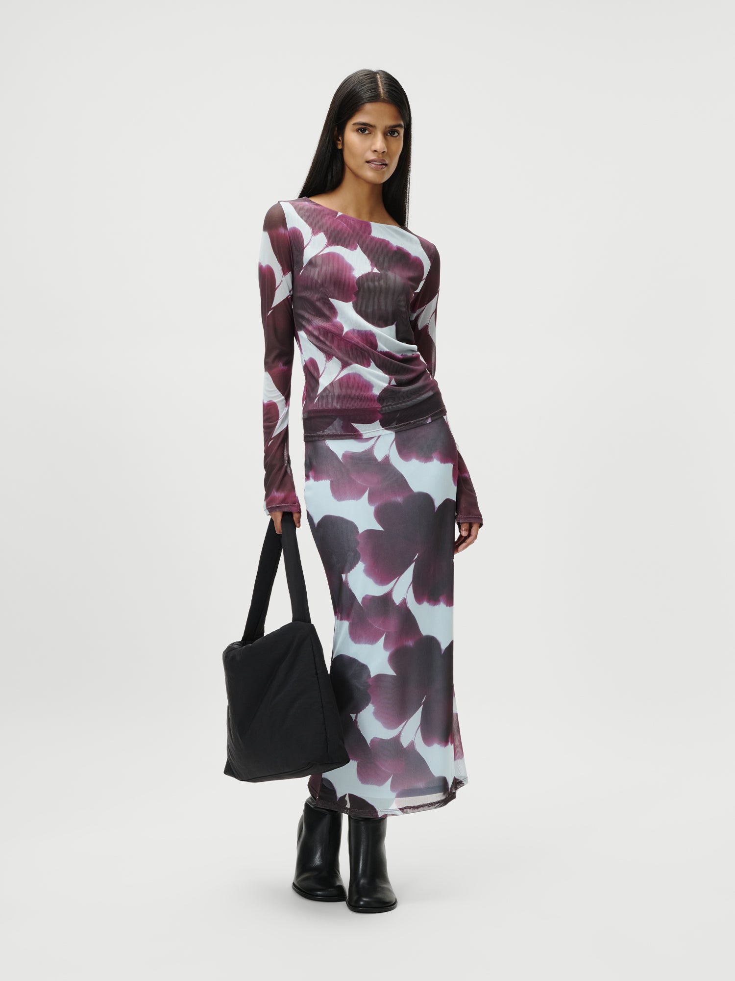 Eine Frau steht vor einem weißen Hintergrund und trägt das Floria Longsleeve von LeGer by Lena Gercke in Größe 36 mit einem auffälligen lila und weißen Blumenmuster. Sie kombiniert es mit schwarzen Stiefeln und trägt eine schwarze Umhängetasche.