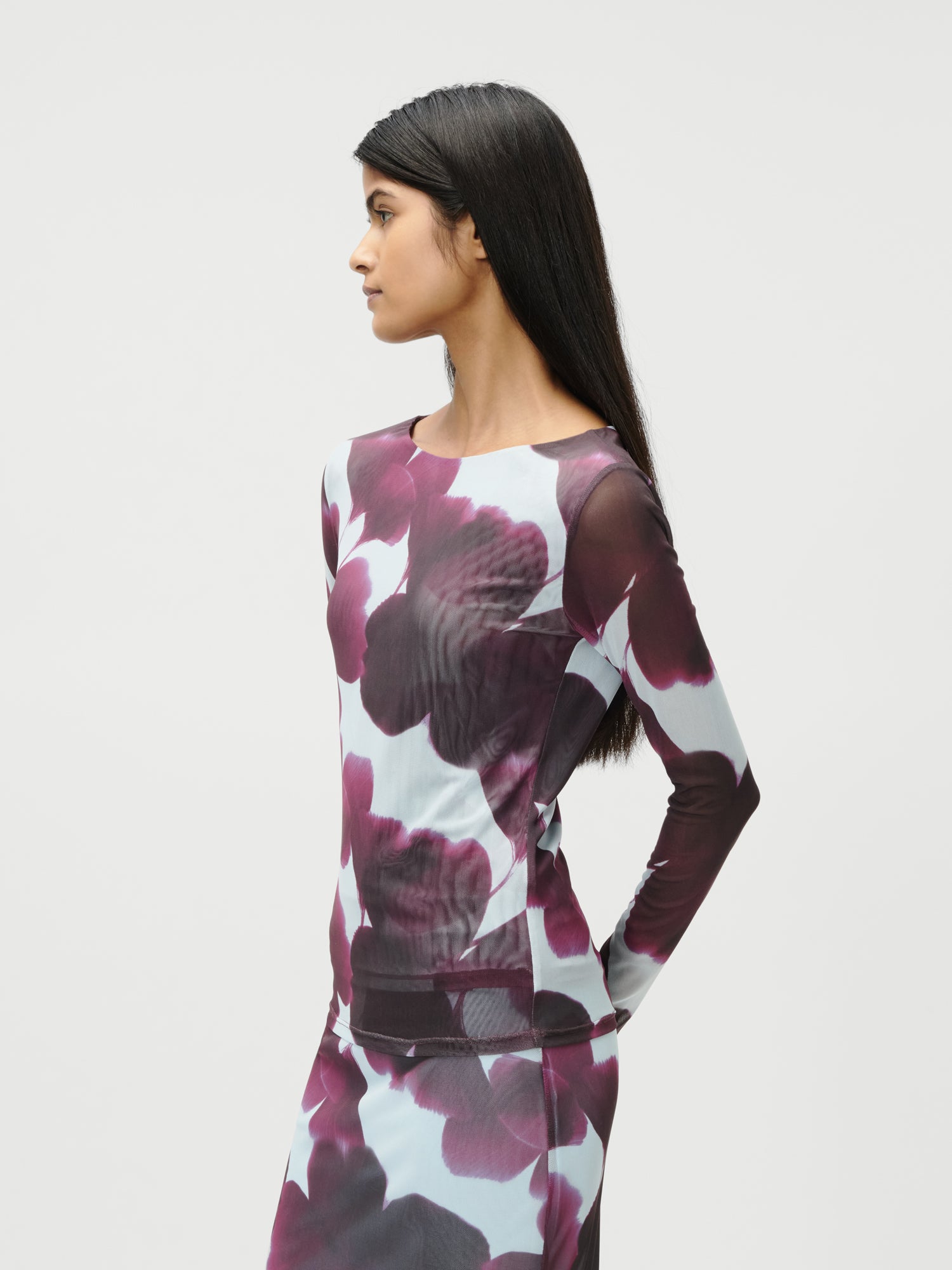 Eine schlanke Frau mit langen dunklen Haaren steht seitlich vor einem weißen Hintergrund. Sie trägt das taillierte Langarmkleid Floria von LeGer by Lena Gercke mit extralangem Schnitt und einem auffälligen lila Blumendruck auf hellem Grund.