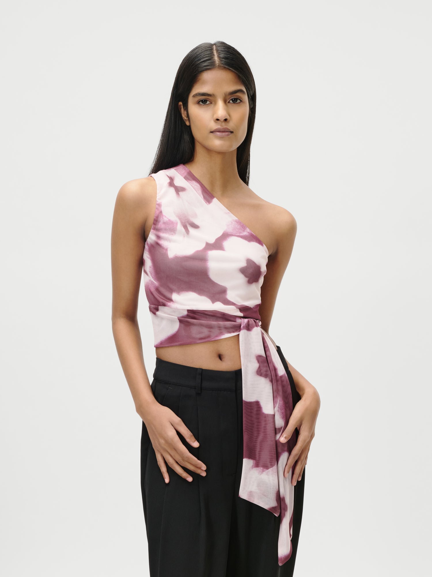 Eine Frau mit langem, glattem schwarzem Haar trägt das LeGer by Lena Gercke Floria Top, ein figurbetontes, ärmelloses rosa-weißes, geblümtes Crop-Top mit seitlicher Bindung und kurzem Schnitt, gepaart mit einer schwarzen Hose mit hoher Taille vor einem schlichten weißen Hintergrund.