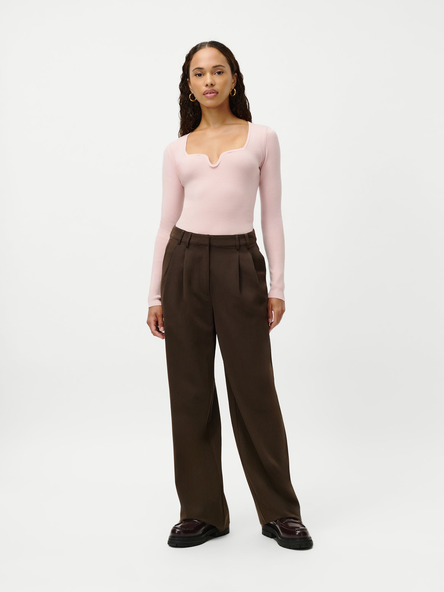 Eine Frau trägt den Guya Jumper von LeGer by Lena Gercke, ein schmal geschnittenes, hellrosa, langärmeliges Oberteil mit quadratischem Ausschnitt, kombiniert mit einer dunkelbraunen, weit geschnittenen Hose und dunklen Schuhen. Sie hat langes, lockiges Haar. Outfit abgebildet in Größe 36.