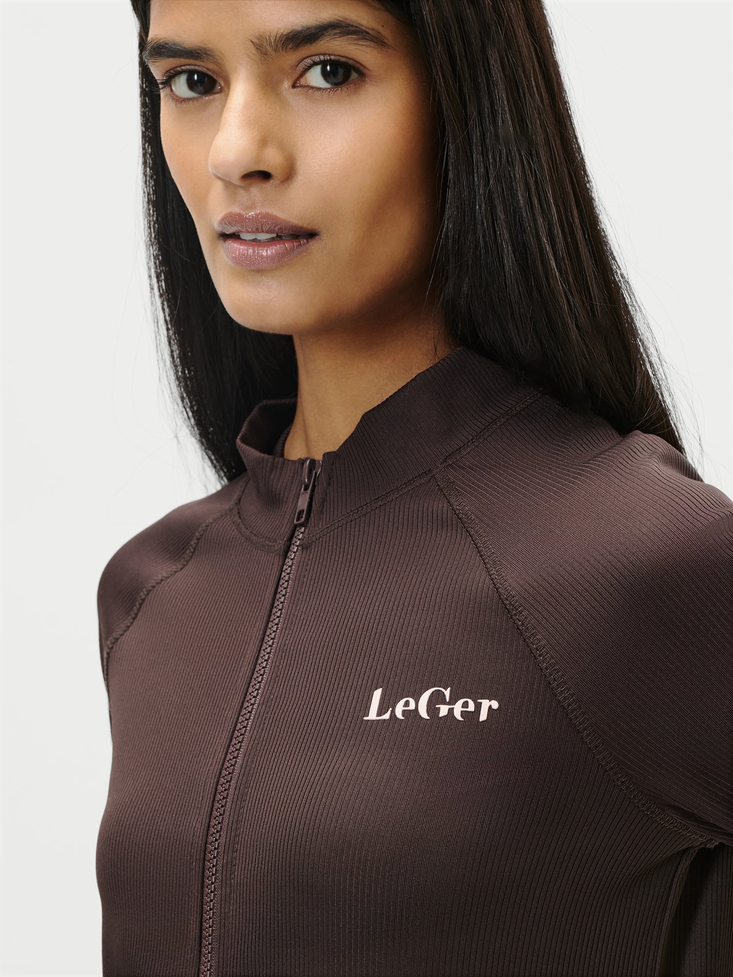 Eine Frau mit langen dunklen Haaren trägt die Simone Jacket von LeGer by Lena Gercke, eine braune Jacke mit Reißverschluss und dem weißen Schriftzug "LeGer" auf der Brust, die vor einem unifarbenen Hintergrund leicht zur Seite schaut.