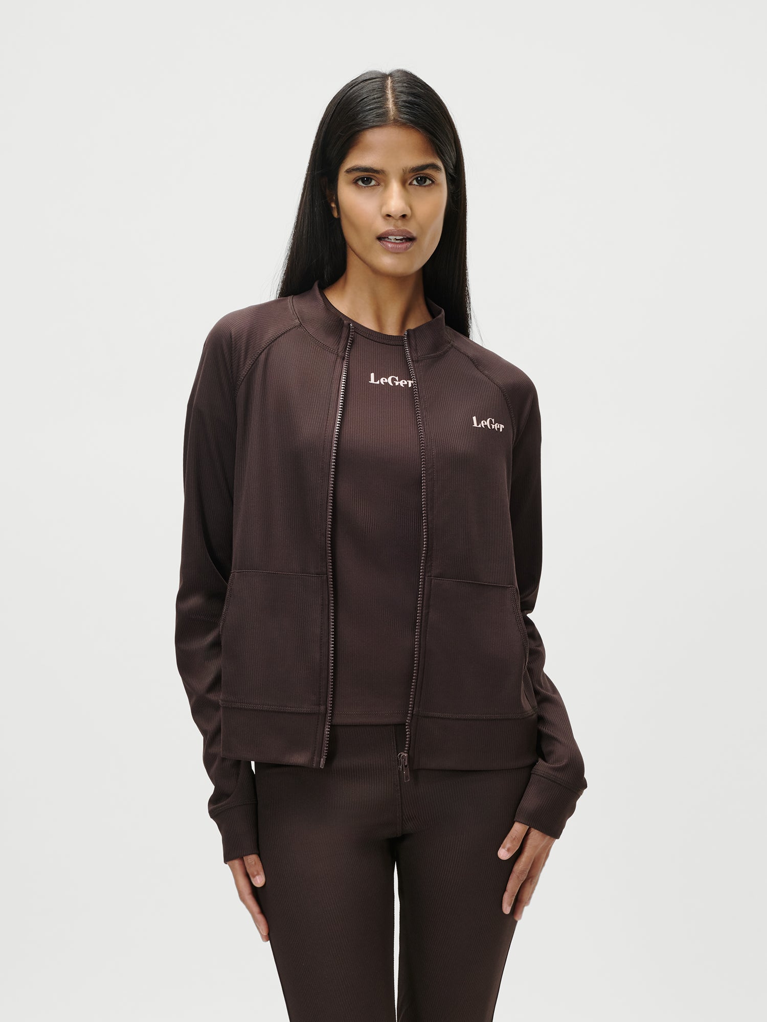Eine Frau mit langen dunklen Haaren steht vor einem weißen Hintergrund und trägt die Jacke Simone von LeGer by Lena Gercke in Dunkelbraun, mit langen Ärmeln und normaler Länge, gepaart mit einer passenden Hose. Auf der Jacke steht "Le Crop", während sie mit den Händen an den Seiten nach vorne blickt.