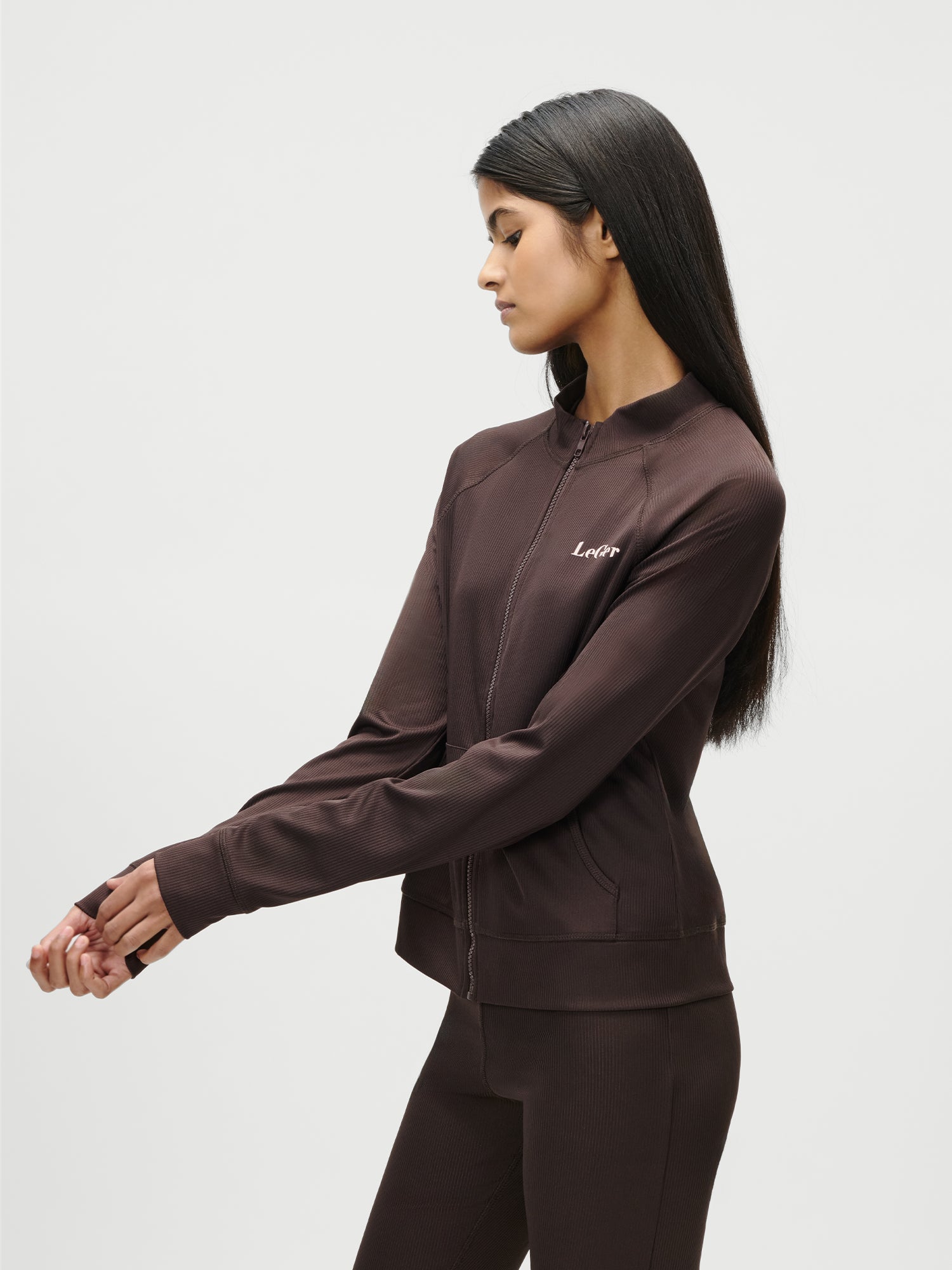 Eine Frau mit langen dunklen Haaren trägt die Simone Jacket von LeGer by Lena Gercke - eine braune Jacke mit Reißverschluss und langen Ärmeln - und dazu eine passende Hose. Sie steht vor einem schlichten weißen Hintergrund und blickt nach links, wobei sie ihre Hände leicht nach vorne streckt.