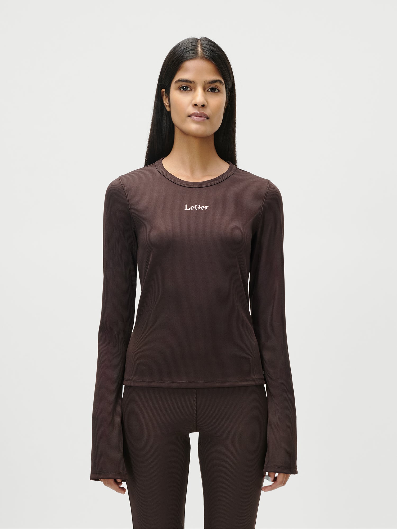Eine Frau mit langen, glatten schwarzen Haaren trägt das taillierte Simone Longsleeve von LeGer by Lena Gercke in Dunkelbraun mit dem Logo auf der Brust und einer dazu passenden Hose, Größe 36, vor einem schlichten weißen Hintergrund.