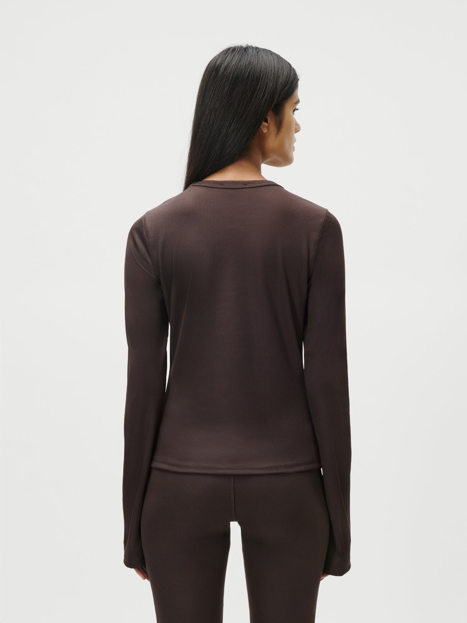 Eine Person mit langen, glatten dunklen Haaren ist von hinten zu sehen, die das LeGer by Lena Gercke Simone Longsleeve in Dunkelbraun mit passender extralanger Hose trägt und vor einem schlichten weißen Hintergrund steht.