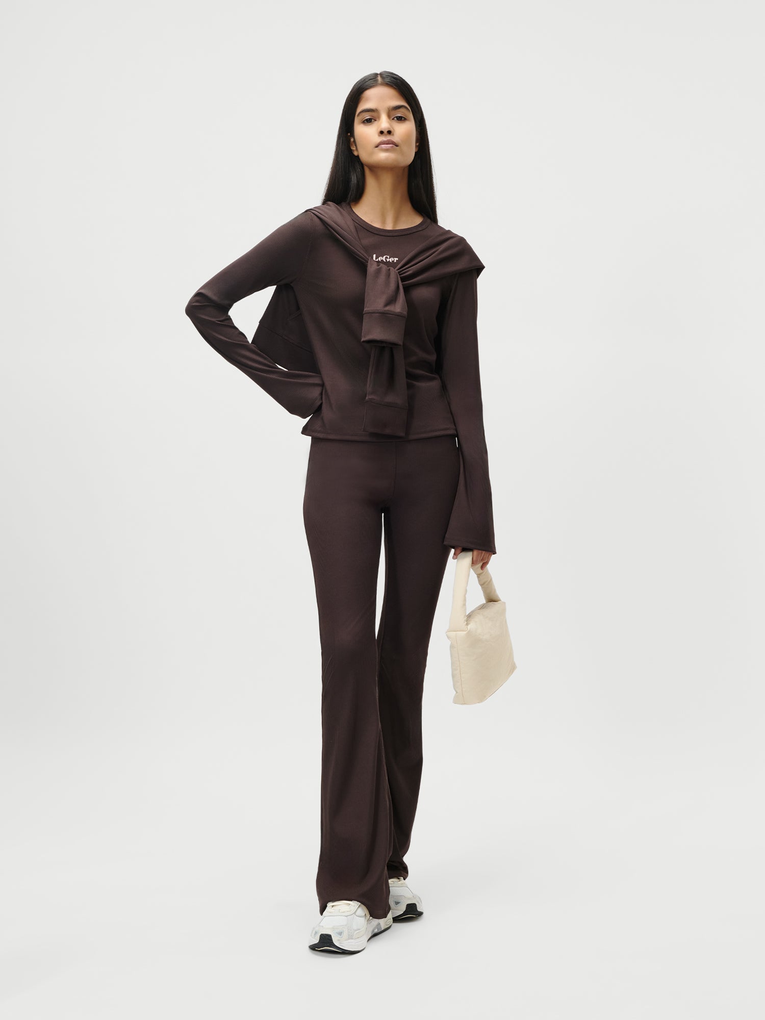 Eine Frau steht vor einem schlichten Hintergrund und trägt das Simone Longsleeve von LeGer by Lena Gercke in Dunkelbraun mit einer passenden extralangen Hose, weißen Turnschuhen, einer kleinen beigen Handtasche und einem ähnlichen braunen Oberteil, das sie sich schick über die Schultern gelegt hat.