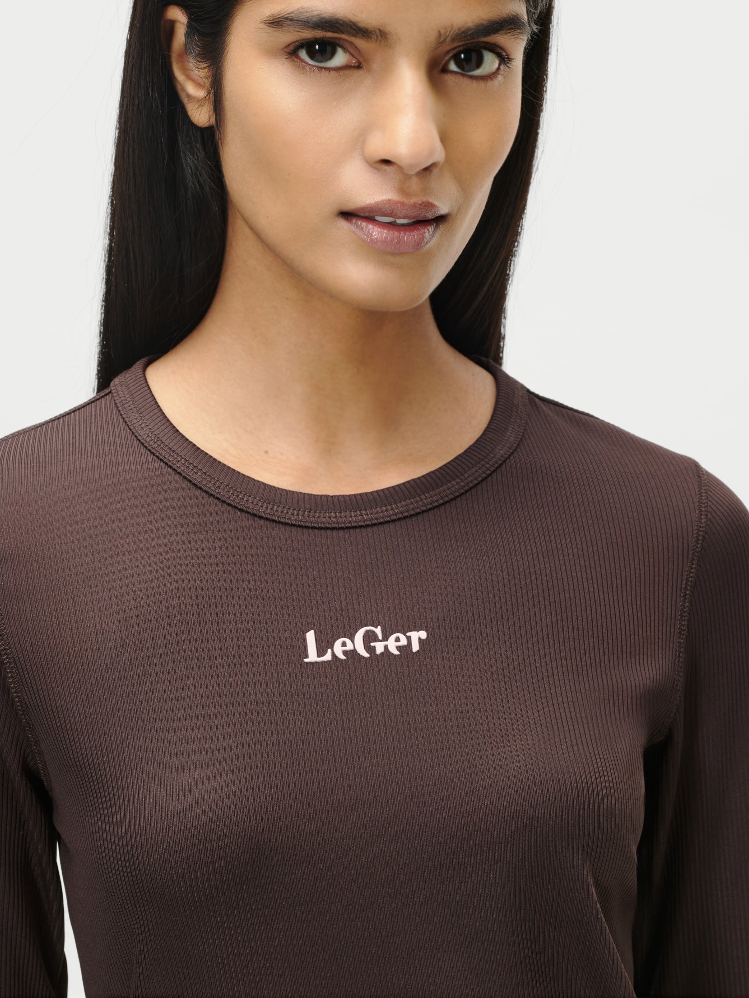 Eine Person mit langen dunklen Haaren trägt das braune gerippte Langarmshirt Simone in Größe 36 von LeGer by Lena Gercke mit dem Schriftzug "LeGer" in weißer Schrift auf der Brust vor einem schlichten hellen Hintergrund.