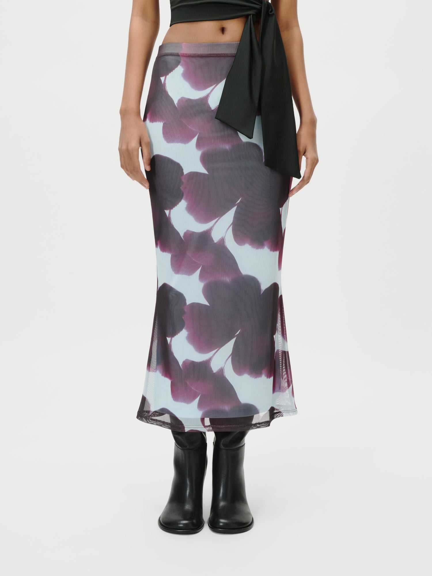 Eine Person trägt den Floria Rock von LeGer by Lena Gercke - einen durchsichtigen, knöchellangen Rock mit auffälligen dunkelvioletten Blumen auf hellem Grund -, kombiniert mit schwarzen Stiefeln und einem schwarzen Oberteil, vor einem schlichten weißen Hintergrund.