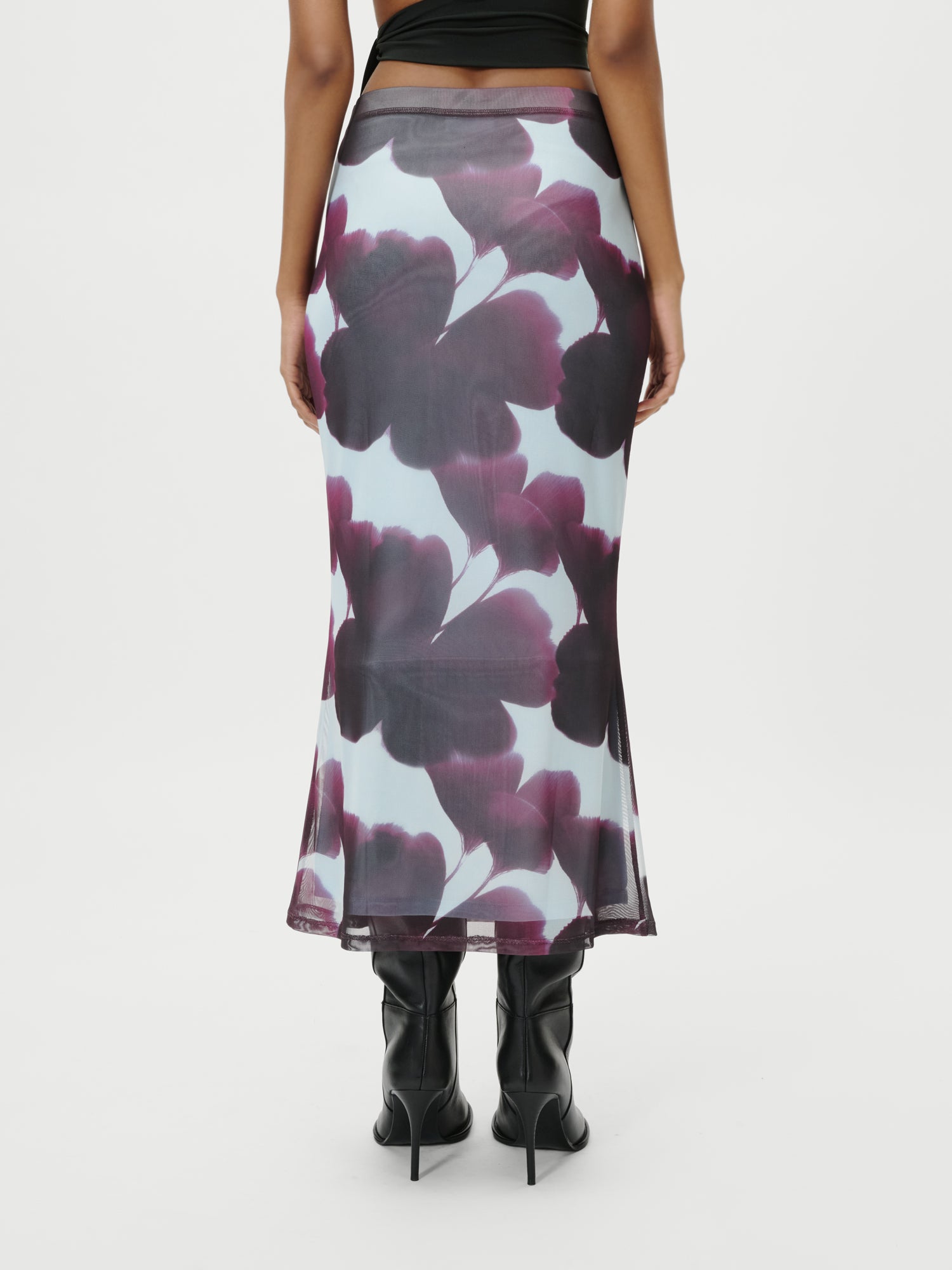 Eine Person, die den Floria Rock von LeGer by Lena Gercke trägt - einen knöchellangen, hochtaillierten, durchsichtigen Rock mit auffälligen lila und dunkelroten Blumendrucken - und dazu schwarze hochhackige Stiefel trägt, fotografiert von hinten auf einem schlichten weißen Hintergrund.