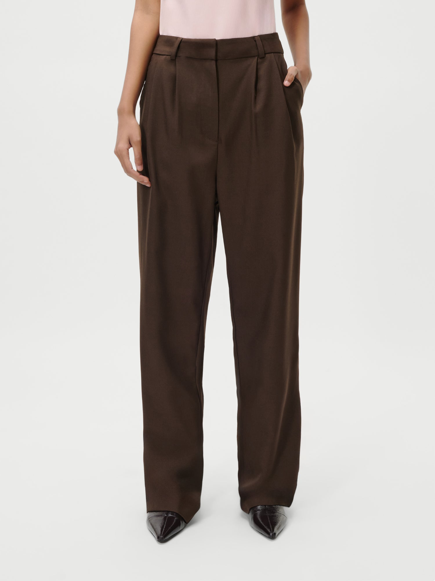 Eine Person steht vor einem unifarbenen weißen Hintergrund und trägt die Jerika Pants von LeGer by Lena Gercke in Größe 36 - eine dunkelbraune, taillierte, locker sitzende Hose mit Bügelfalten und Taschen auf der Vorderseite -, dazu ein hellrosa Oberteil und schwarze Schuhe mit spitzen Zehen.