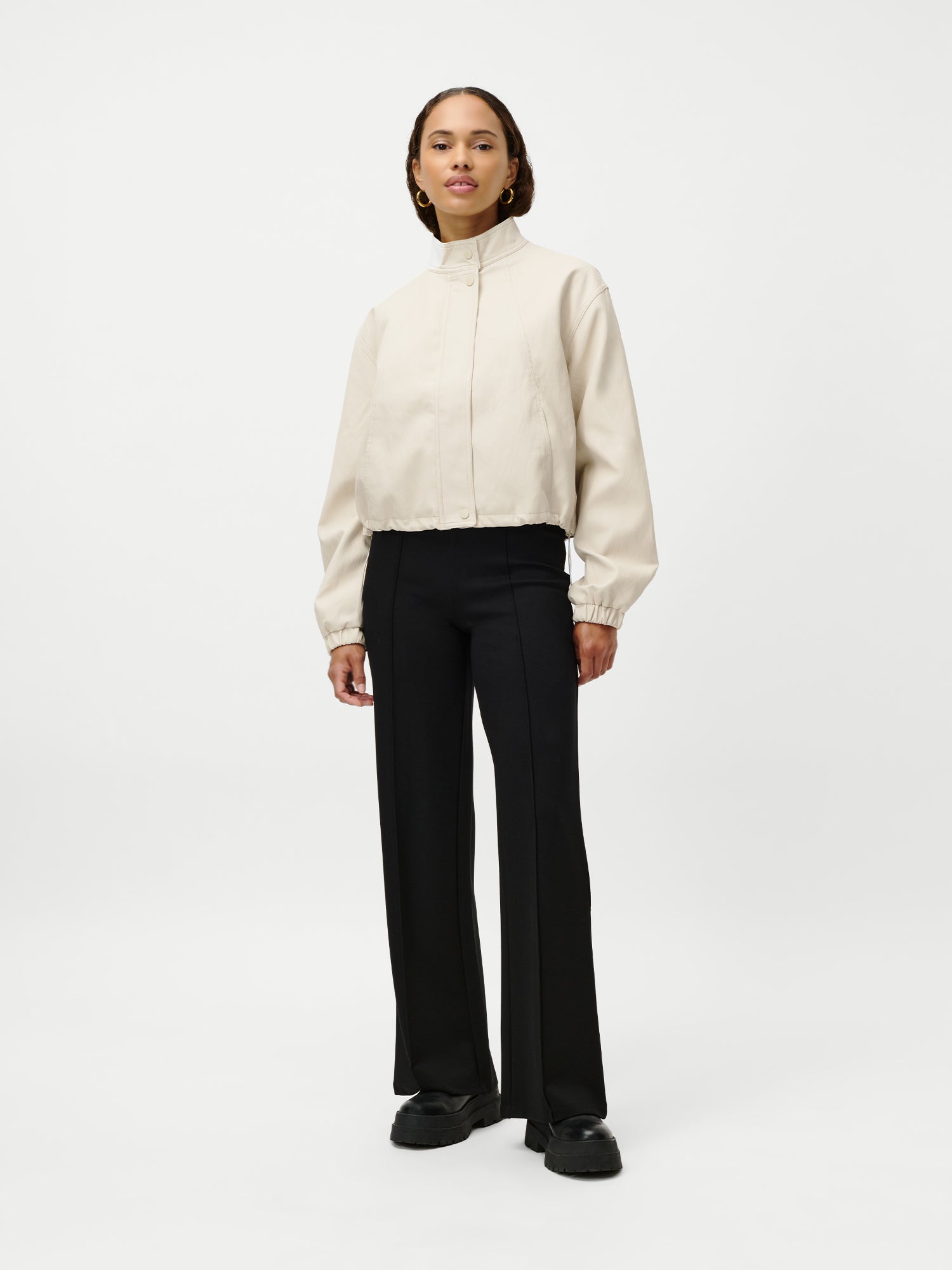 Eine Frau steht vor einem schlichten weißen Hintergrund und trägt eine hellbeige Janet Jacket von LeGer by Lena Gercke in Größe 36, dazu eine schwarze Hose mit weitem Bein und schwarze Schuhe. Sie blickt nach vorne und hat einen neutralen Gesichtsausdruck.