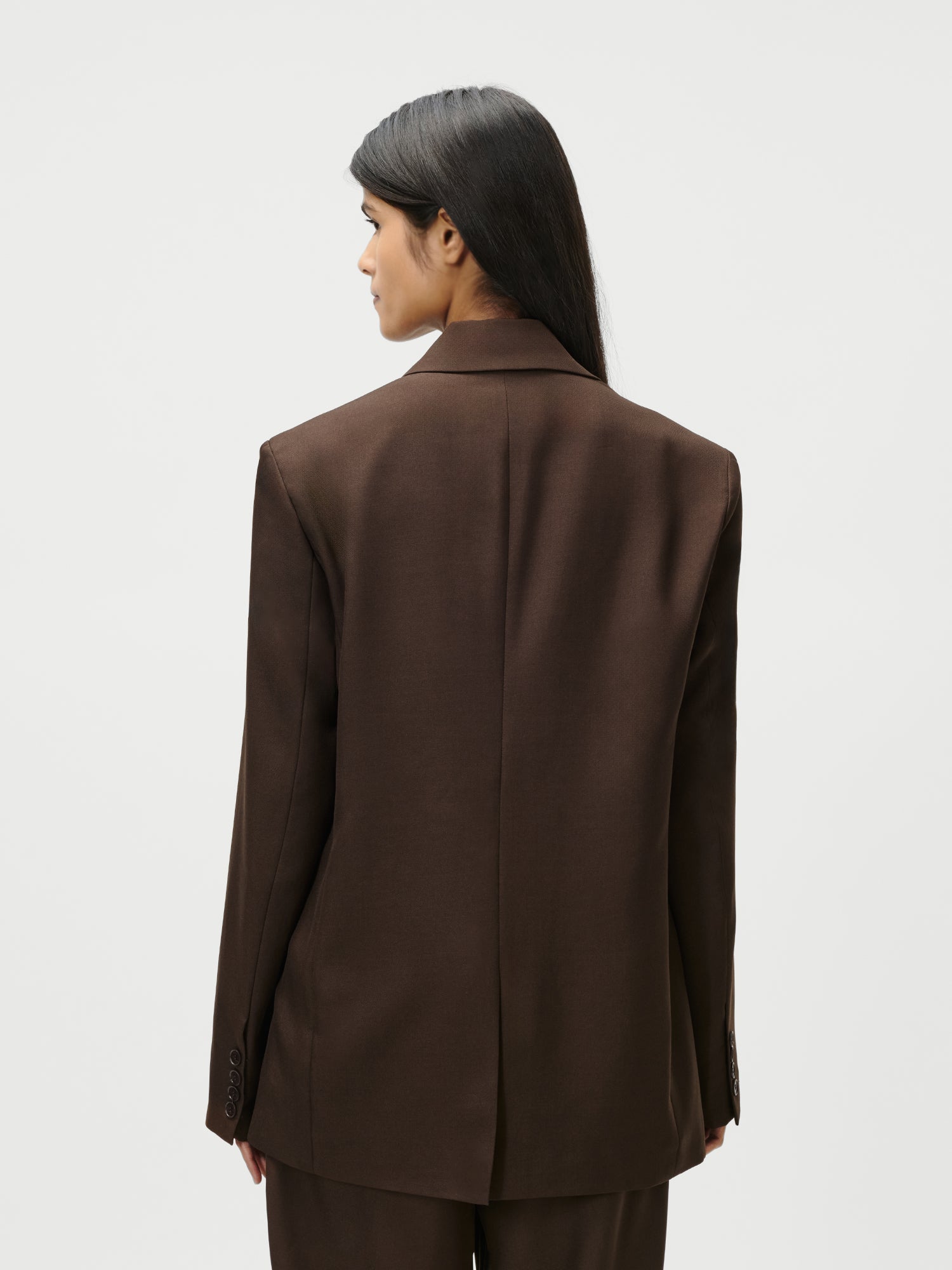 Eine Person mit langen dunklen Haaren, die den Jerika Blazer von LeGer by Lena Gercke in Größe 36, normale Länge und Langarm mit passender Hose trägt, steht von der Kamera abgewandt vor einem schlichten hellen Hintergrund.