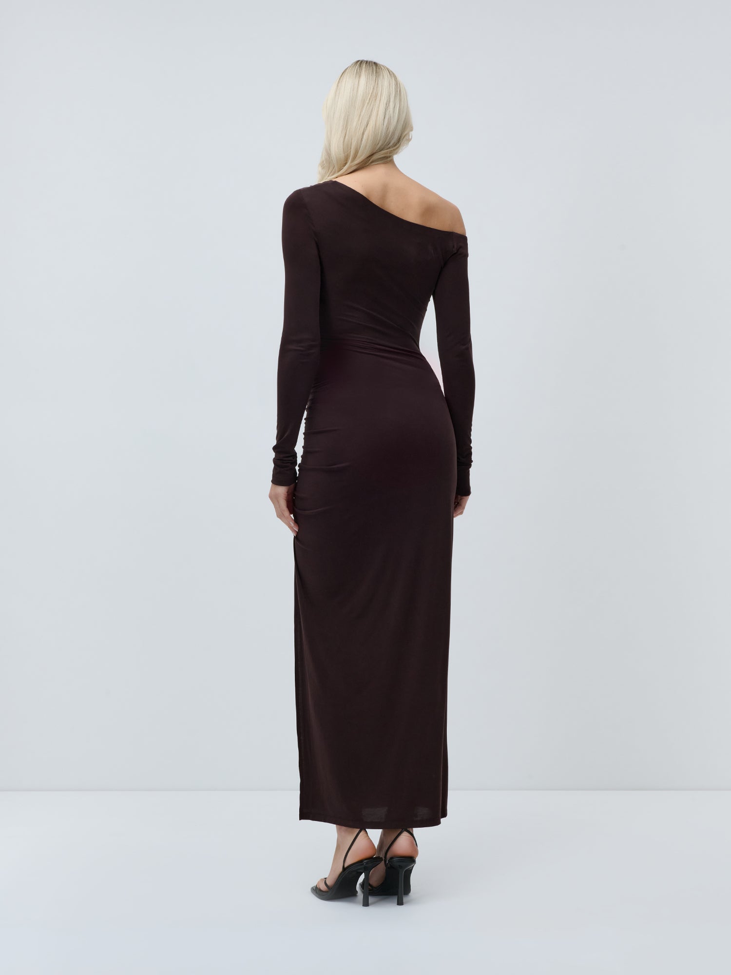 Eine Frau mit langen blonden Haaren präsentiert das schmal geschnittene, dunkelbraune „Adline“-Kleid von LeGer by Lena Gercke in Kombination mit schwarzen Sandalen mit Absatz und steht dabei mit dem Rücken zur Kamera vor einem schlichten, hellen Hintergrund.