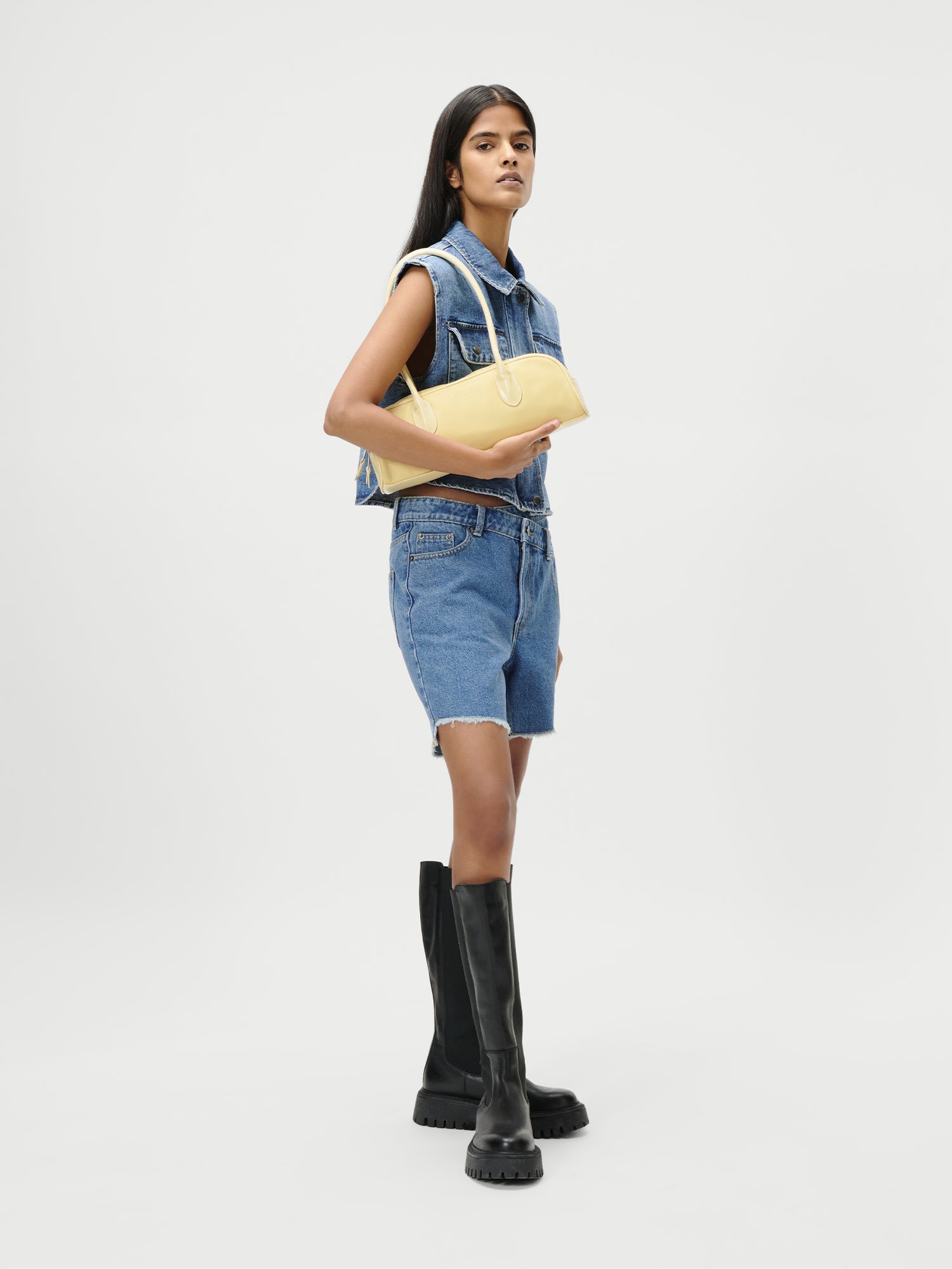 Eine Frau posiert vor weißem Hintergrund in einer ärmellosen Jeansjacke, Jeansshorts und schwarzen kniehohen Stiefeln und hält die Tasche „Frauke“ von LeGer by Lena Gercke in der Hand – ein auffälliges Accessoire, das sich perfekt für schicke Frühlingslooks eignet.