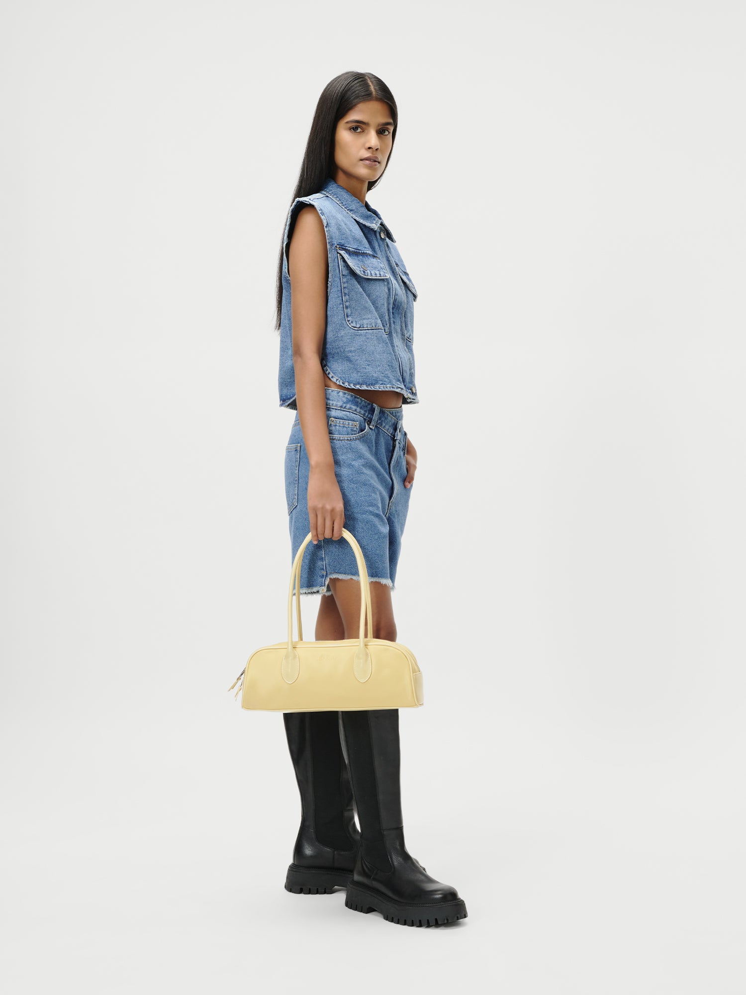 Eine Frau mit langen, dunklen Haaren trägt eine ärmellose Jeansweste, dazu passende Shorts und schwarze kniehohe Stiefel. Sie steht vor einem schlichten weißen Hintergrund und hält die LeGer by Lena Gercke Tasche im Stil „Frauke“ als Blickfang in der Hand.
