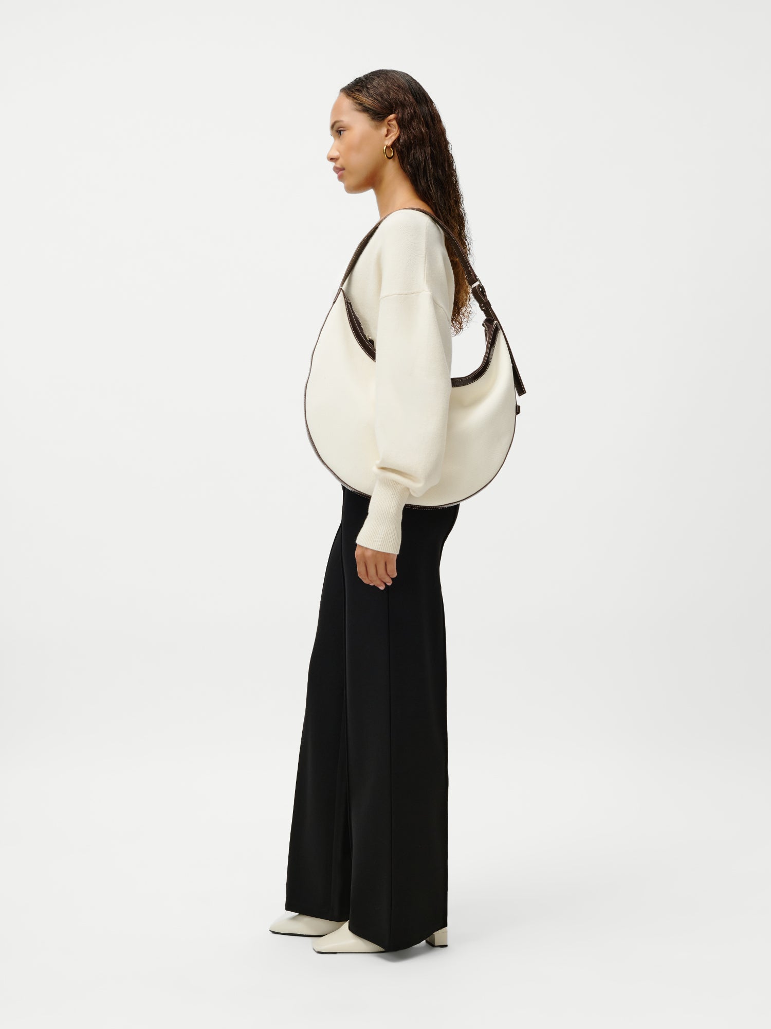 Eine Frau steht seitlich im Bild und trägt einen cremefarbenen Pullover, eine schwarze Hose mit weitem Bein und cremefarbene Schuhe. In der Hand hält sie die Tasche „Gisa“ von LeGer by Lena Gercke – ein elegantes Accessoire mit dunklem Besatz. Der Hintergrund ist schlicht weiß.