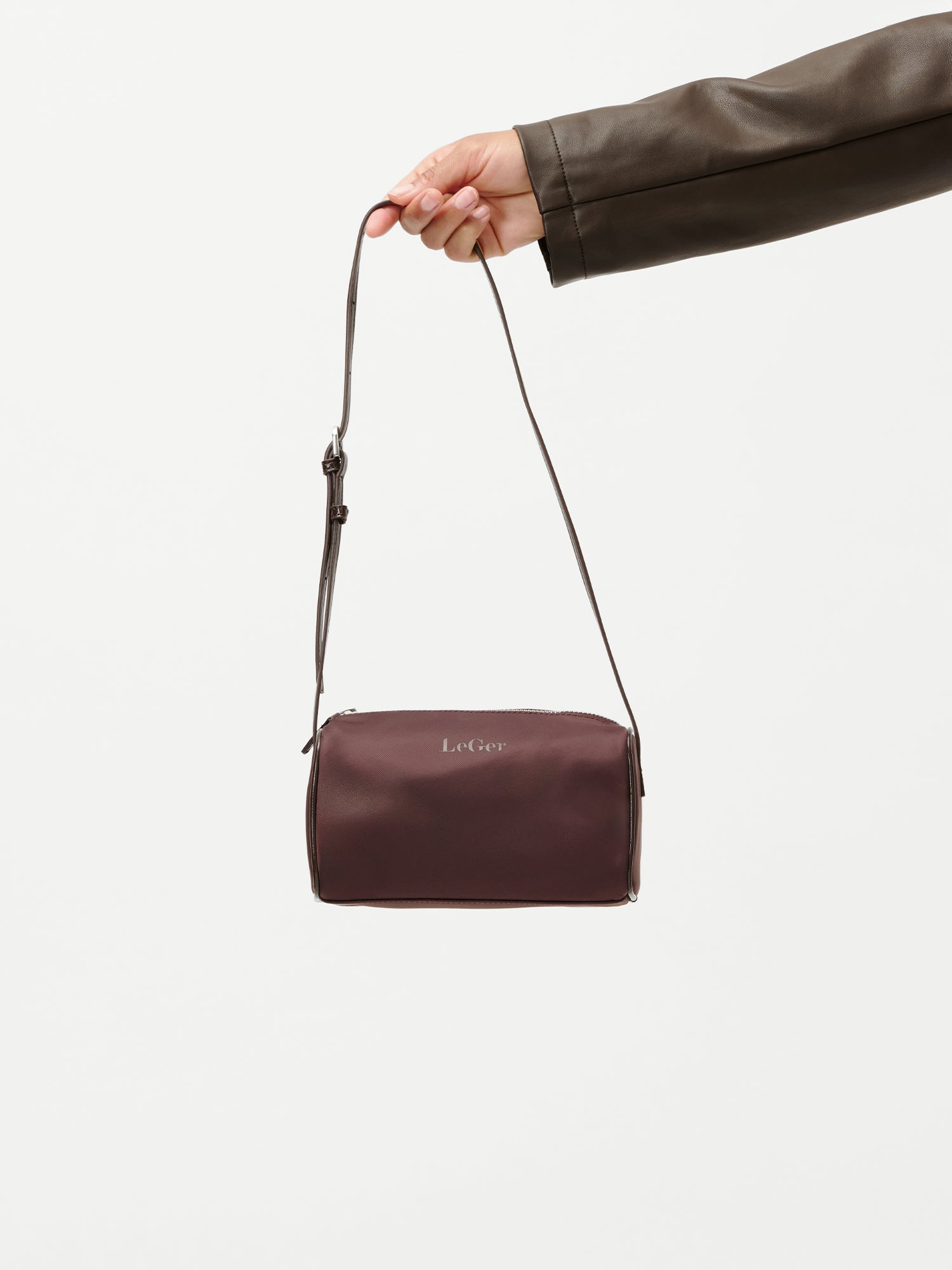 Eine Hand in einem braunen Lederhandschuh hält die „Georgia“-Tasche von LeGer by Lena Gercke – eine kleine, rechteckige, weinrote Umhängetasche mit verstellbarem Riemen und dem Schriftzug „LeGer“ auf der Vorderseite – vor einem schlichten weißen Hintergrund.