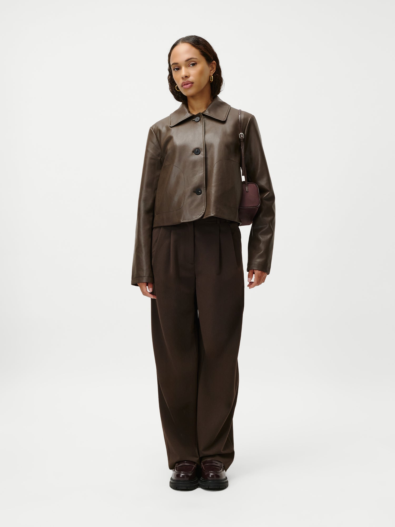 Eine Frau blickt nach vorne; sie trägt eine braune Lederjacke, eine weite braune Hose und dunkle Schuhe und hat die „Georgia“-Tasche von LeGer by Lena Gercke dabei – ein schickes Accessoire, das sich deutlich vom schlichten weißen Hintergrund abhebt.