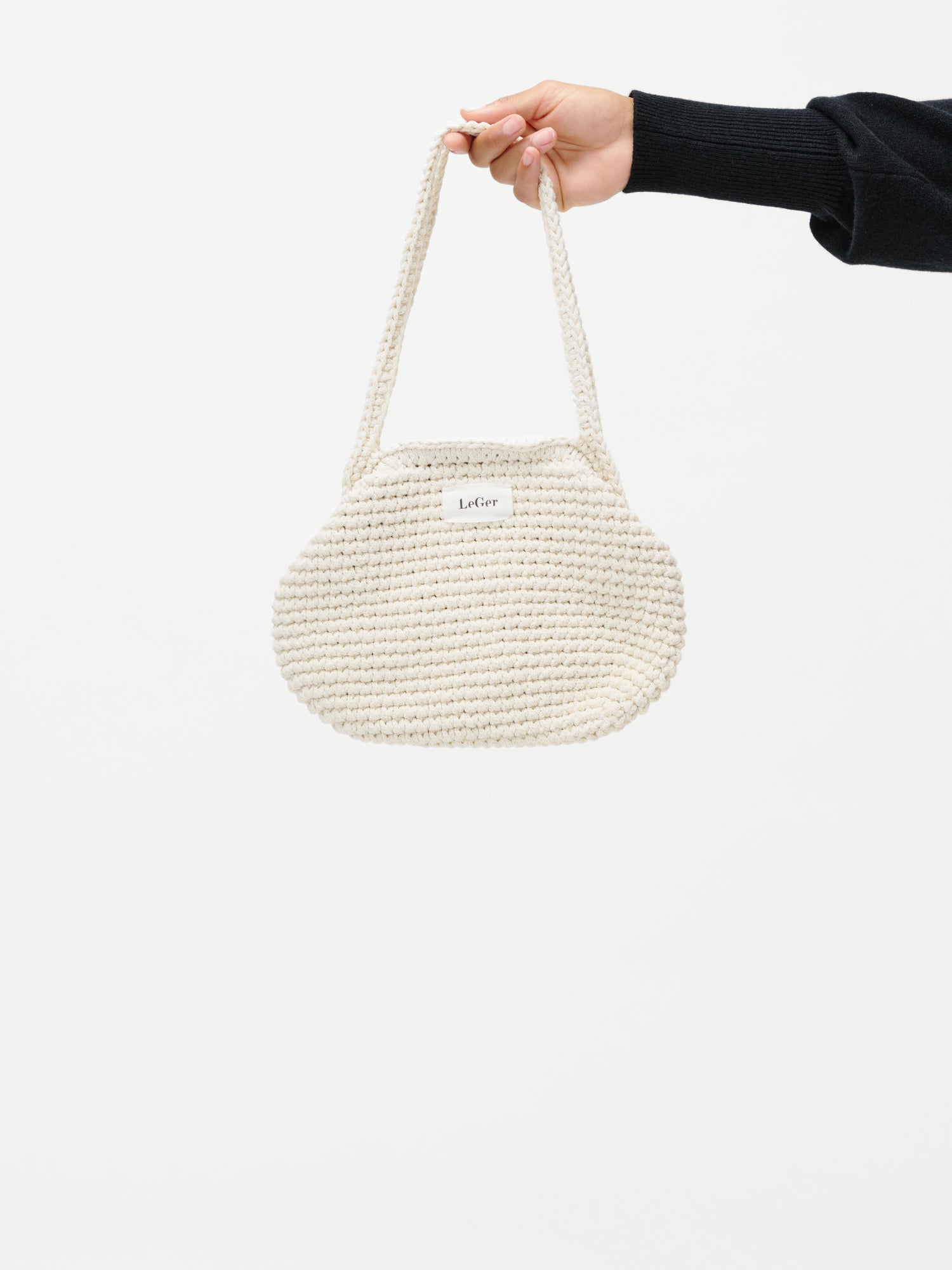 Eine Hand in einem schwarzen Ärmel hält die runde, cremefarbene Perle Bag von LeGer by Lena Gercke, die mit einem kurzen Riemen versehen ist, vor einem schlichten weißen Hintergrund.