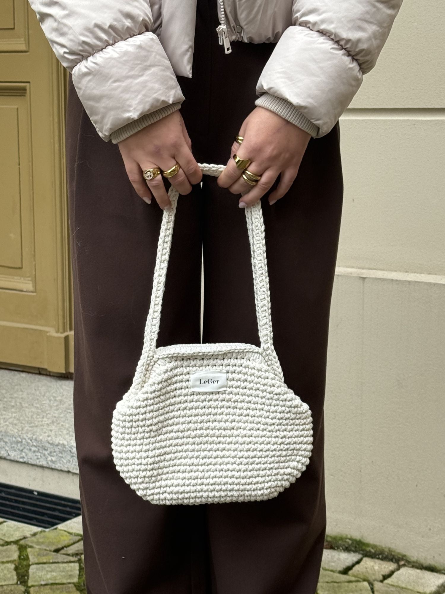 Eine Person in einem hellen Puff und einer braunen Hose hält die LeGer by Lena Gercke Perle Bag im ecrufarbenen Häkel-Stil mit goldenen Ringen in der Hand. Eine braune Tür und ein Steinpflaster bilden den Hintergrund.