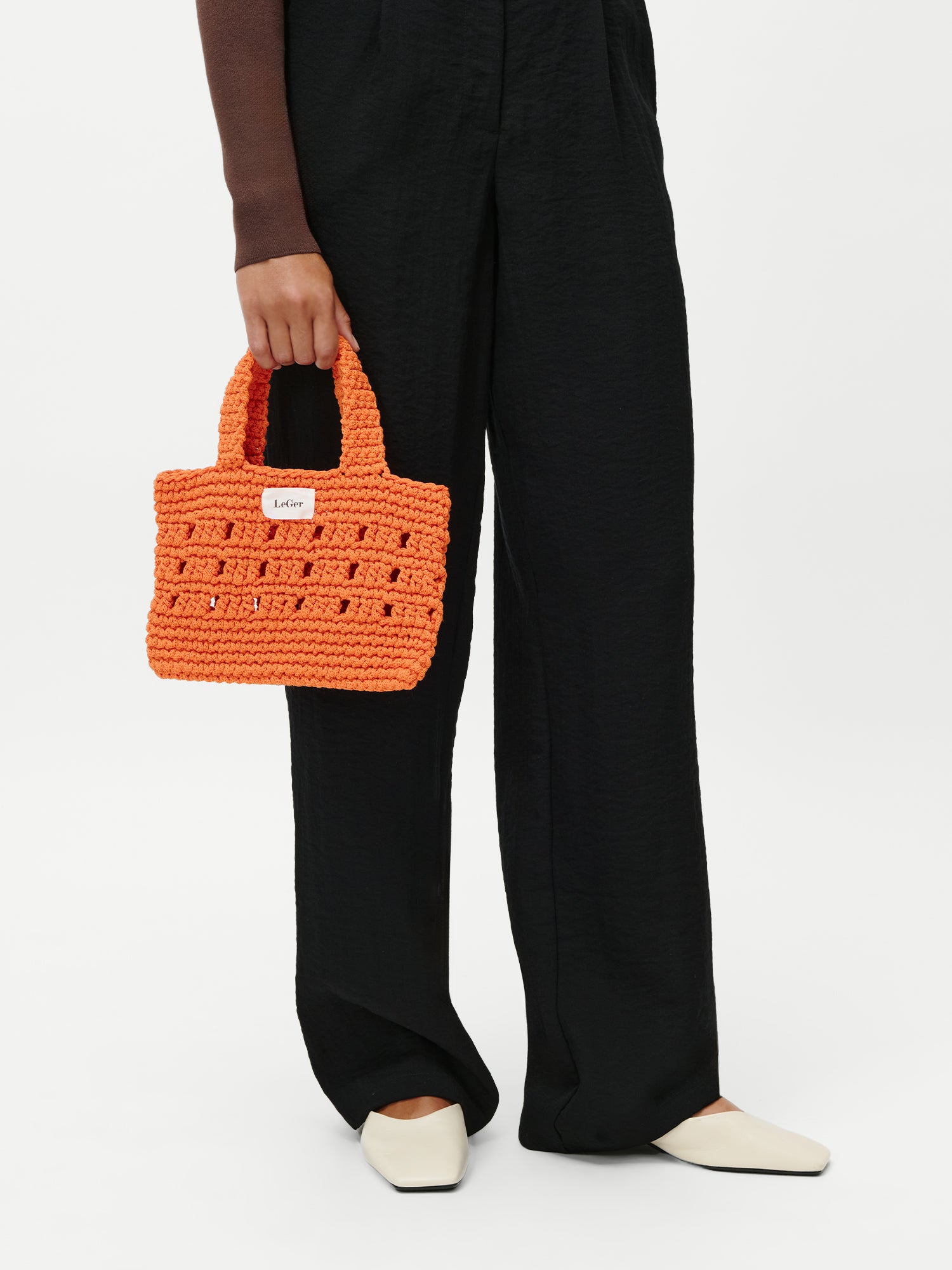 Eine Person hält die stilvolle „Valerie Bag“ von LeGer by Lena Gercke in der Hand – ein kleines, orangefarbenes Häkelmodell mit kurzen Henkeln und einem weißen Etikett – und trägt dazu eine schwarze Hose und cremefarbene Schuhe.