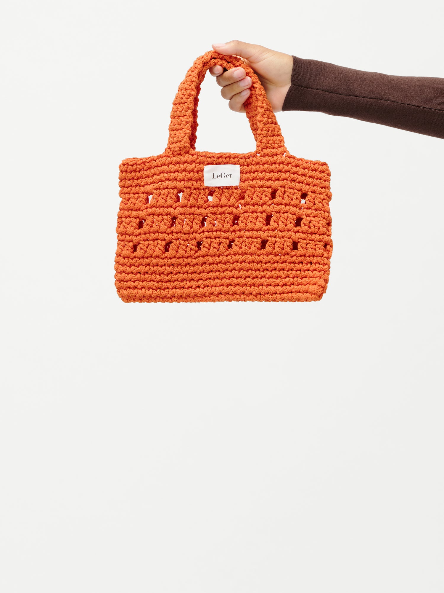 Eine Hand hält die orangefarbene „Valerie“-Tasche von LeGer by Lena Gercke, die sich durch eine feine Häkelstruktur, zwei Henkel und ein kleines weißes Etikett auf der Vorderseite auszeichnet, das sich von einem schlichten weißen Hintergrund abhebt.