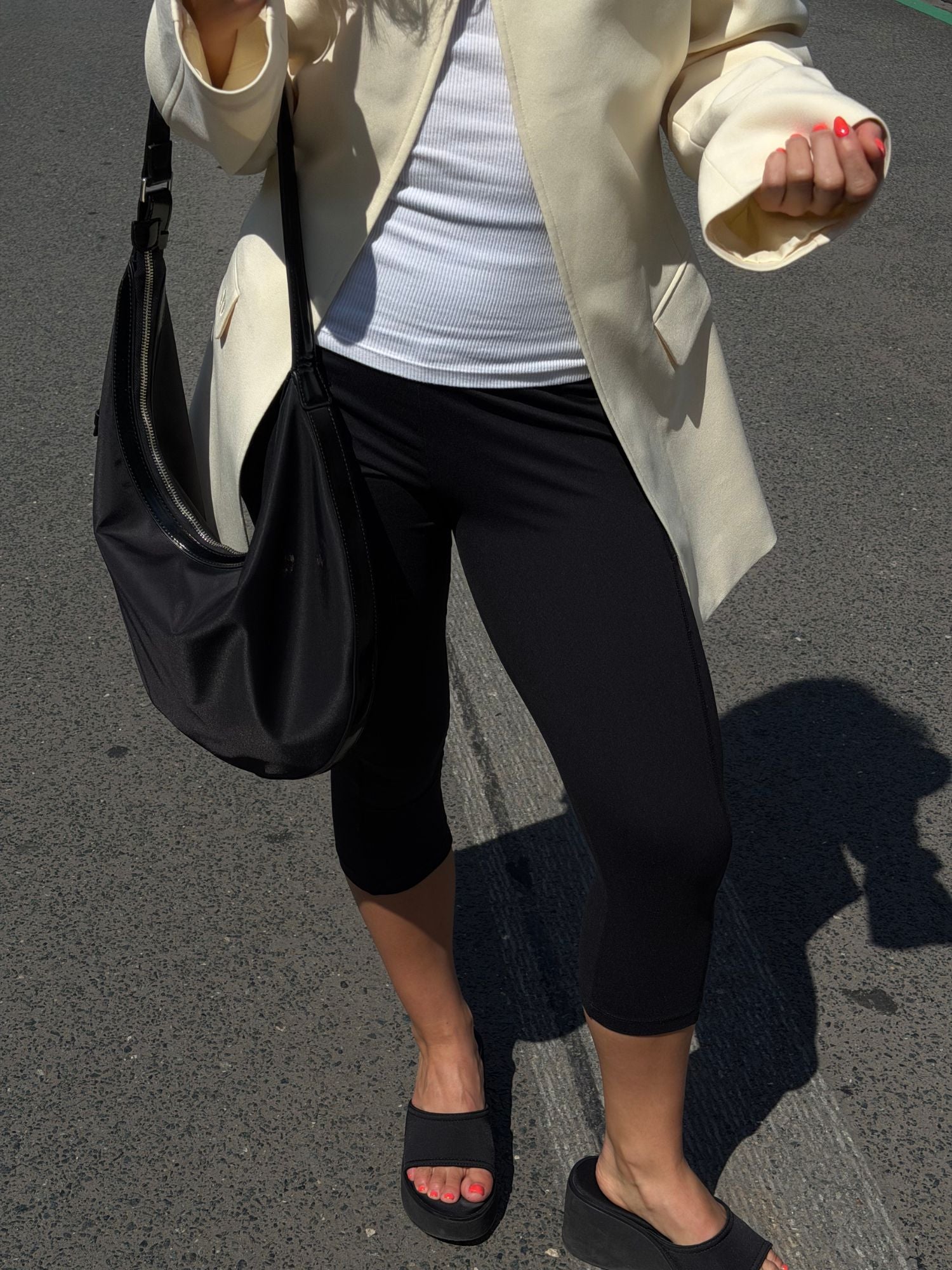 Eine Frau steht auf dem Bürgersteig und trägt einen weißen Blazer, ein weißes Oberteil, schwarze Capri-Leggings und schwarze Pantoletten. Sie hält die elegante „Sarina“-Tasche von LeGer by Lena Gercke in ihrer klassischen Halbmondform in der Hand. Ihre roten Fingernägel und Zehennägel runden dieses schicke LeGer-Outfit perfekt ab.