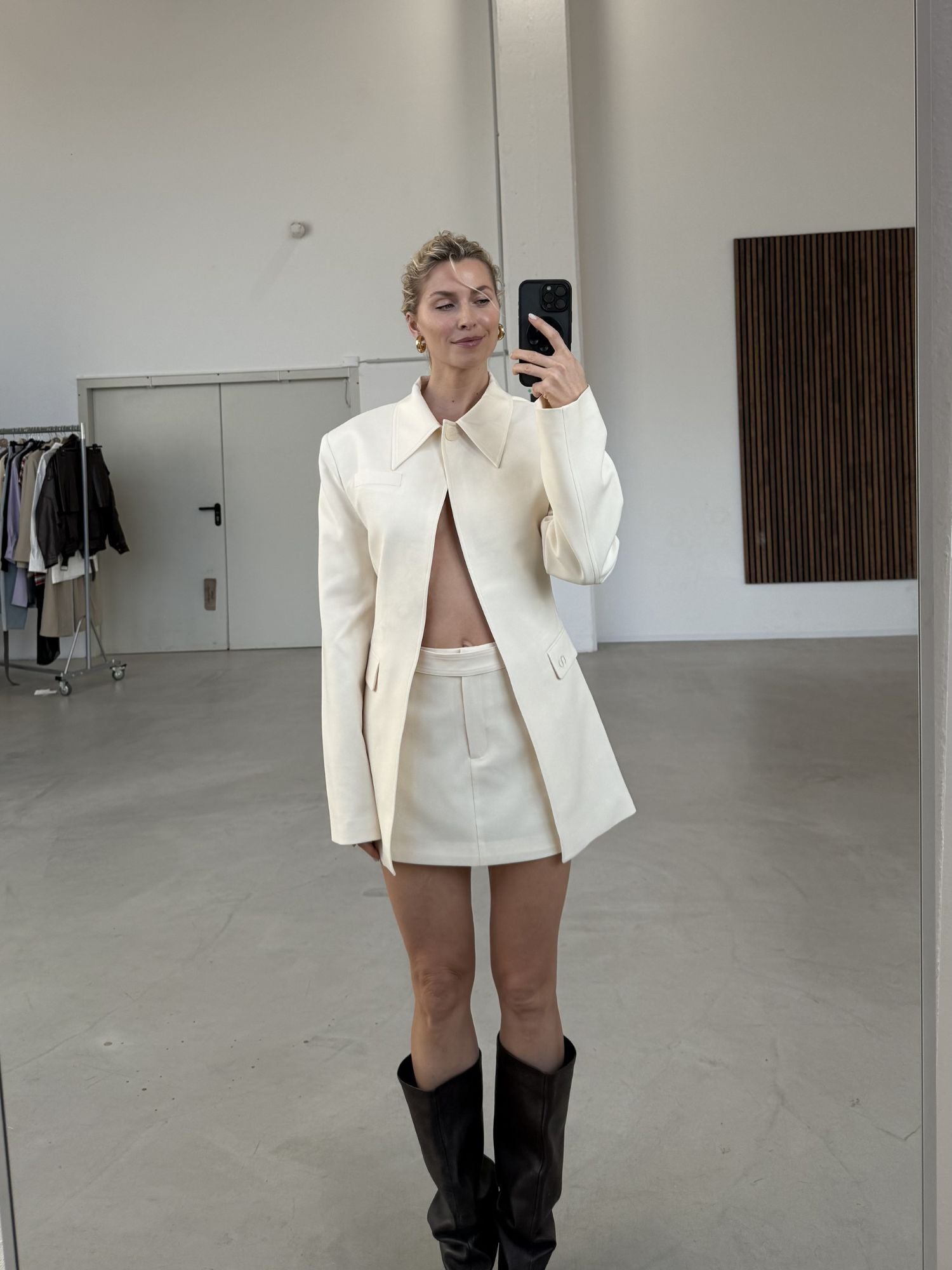 Eine Person steht in einem minimalistischen, modernen Raum und macht ein Spiegel-Selfie. Sie trägt den LeGer by Lena Gercke Cim Blazer mit einem passenden Minirock und dunklen kniehohen Stiefeln. Im Hintergrund ist ein Kleiderständer zu sehen.