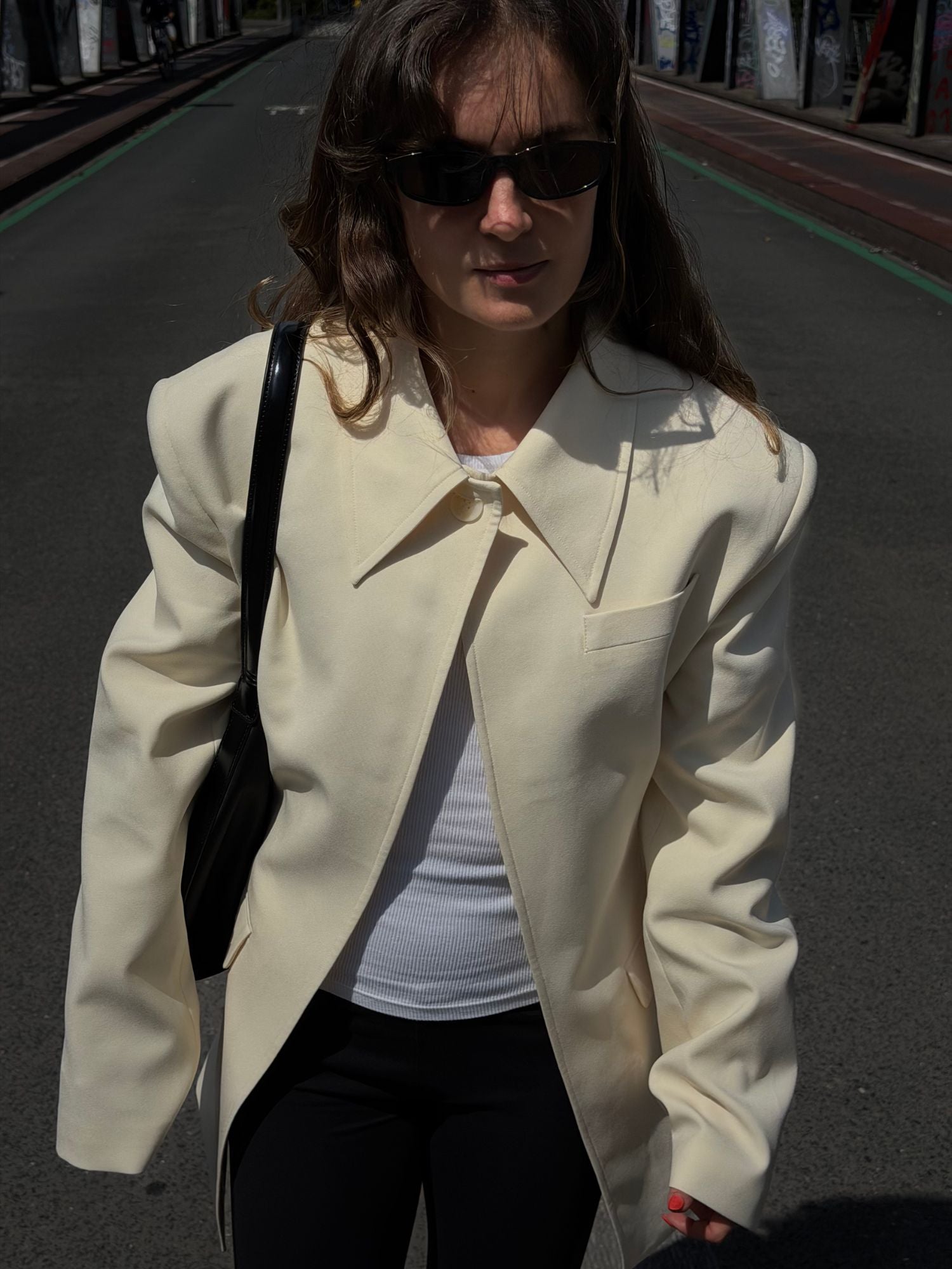 Eine Person, die den cremefarbenen „Cim“-Blazer mit Hemdkragen von LeGer by Lena Gercke trägt, kombiniert mit einem weißen Oberteil, einer schwarzen Hose und einer Sonnenbrille, geht eine Straße entlang, die von mit Graffiti bedeckten Wänden gesäumt ist, und präsentiert dabei elegante Damenmode.