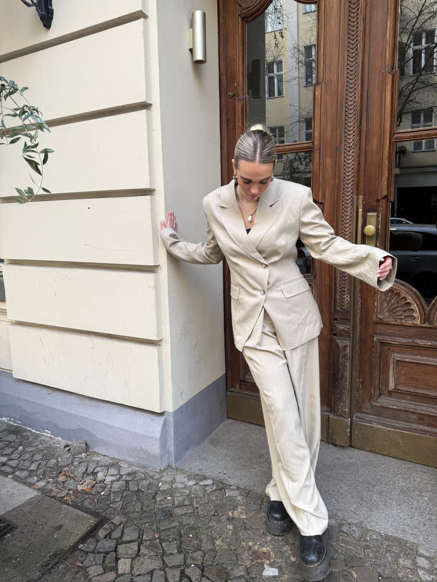 Eine Person mit zurückgebundenen blonden Haaren trägt den beigefarbenen Raquel Blazer von LeGer by Lena Gercke mit einer asymmetrischen Verschlussleiste und schwarzen Schuhen. Sie steht in der Nähe einer Holztür und lehnt sich an ein cremefarbenes Gebäude mit Kopfsteinpflaster.