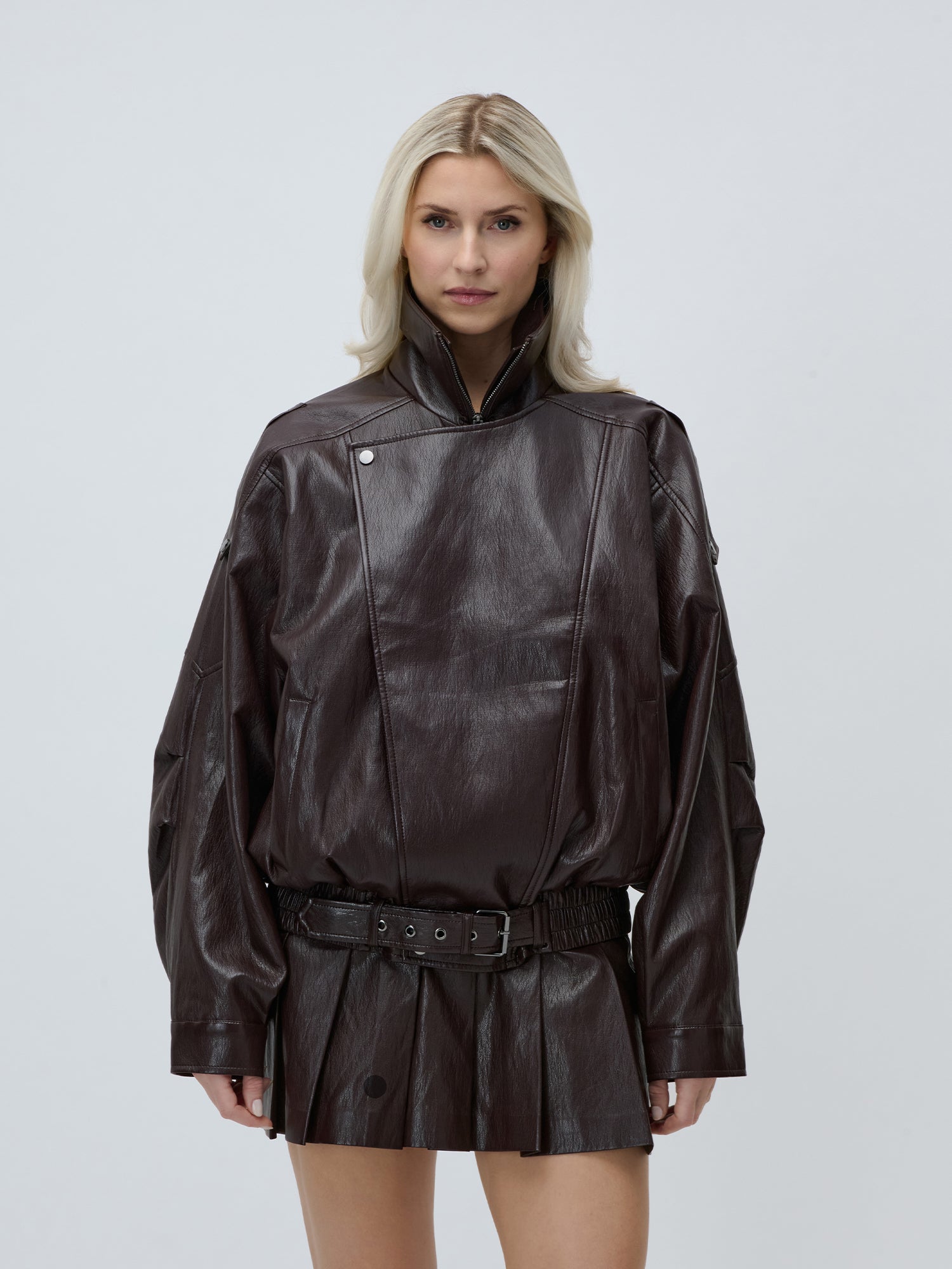Eine blonde Frau modelliert die LeGer by Lena Gercke Jessa Jacket, eine übergroße dunkelbraune Bikerjacke aus Kunstleder mit hohem Kragen, gepaart mit einem passenden Faltenminirock. Sie steht vor einem unifarbenen, hellgrauen Hintergrund und präsentiert einen urbanen Stil.