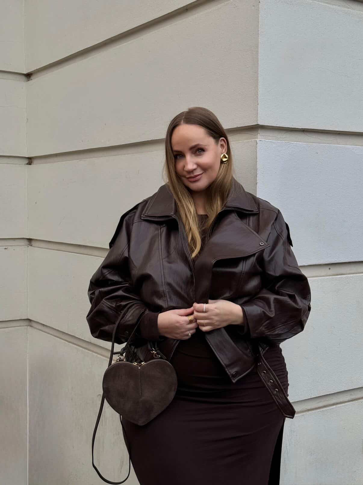 Eine Frau steht vor einer hellen Wand und trägt die dunkelbraune Jessa Jacket von LeGer by Lena Gercke mit einem passenden Kleid und goldenen Reifohrringen. In der Hand hält sie eine braune Handtasche in Herzform, die ihr einen mühelos schicken urbanen Look verleiht, während sie sanft in die Kamera lächelt.