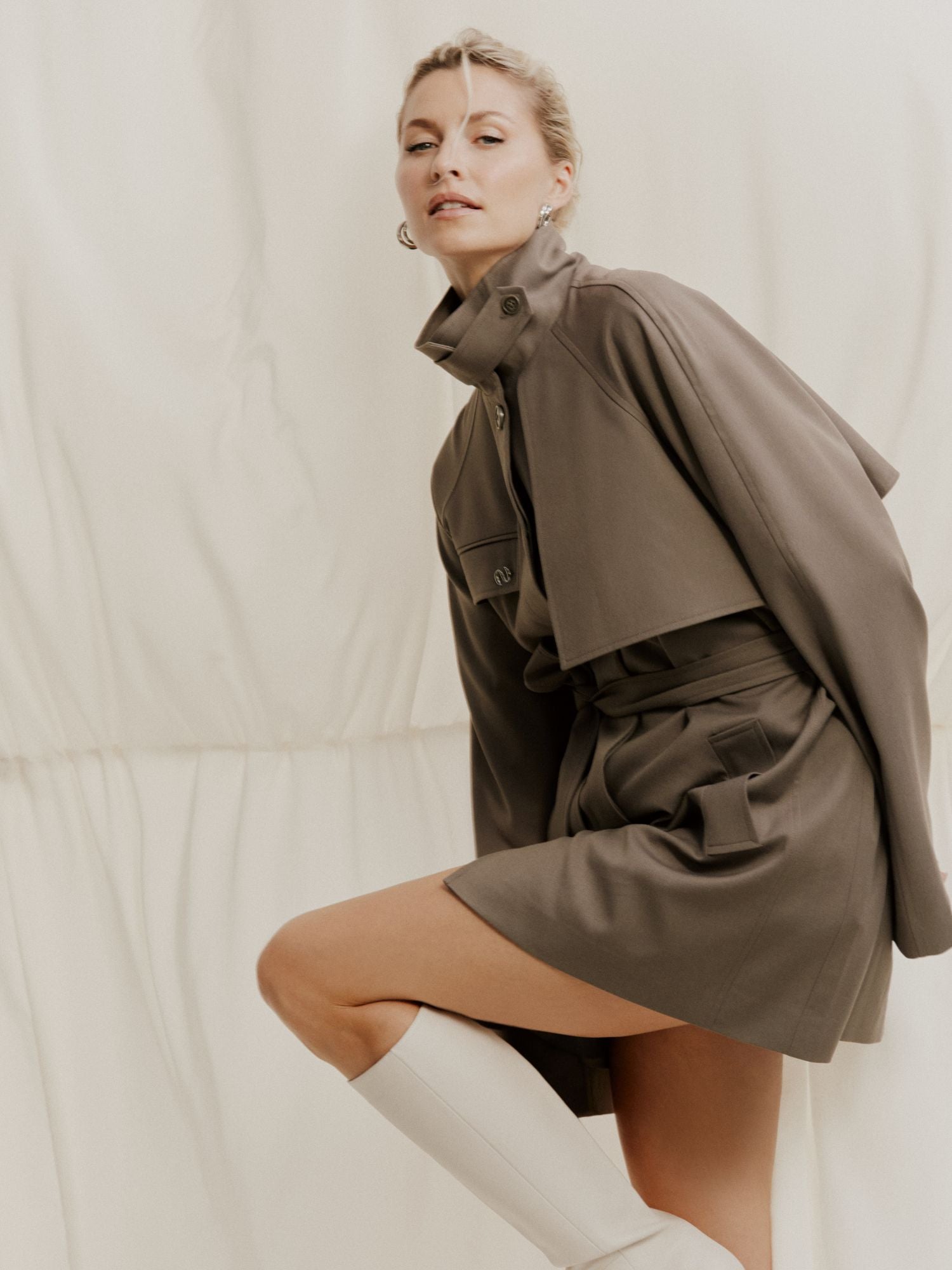Eine Frau posiert selbstbewusst in der Fee Jacket von LeGer by Lena Gercke, einer taupefarbenen Jacke im Trenchcoat-Stil mit Cape-Detail und passenden Shorts, die mit weißen kniehohen Stiefeln vor einem zart cremefarbenen Hintergrund kombiniert wird.