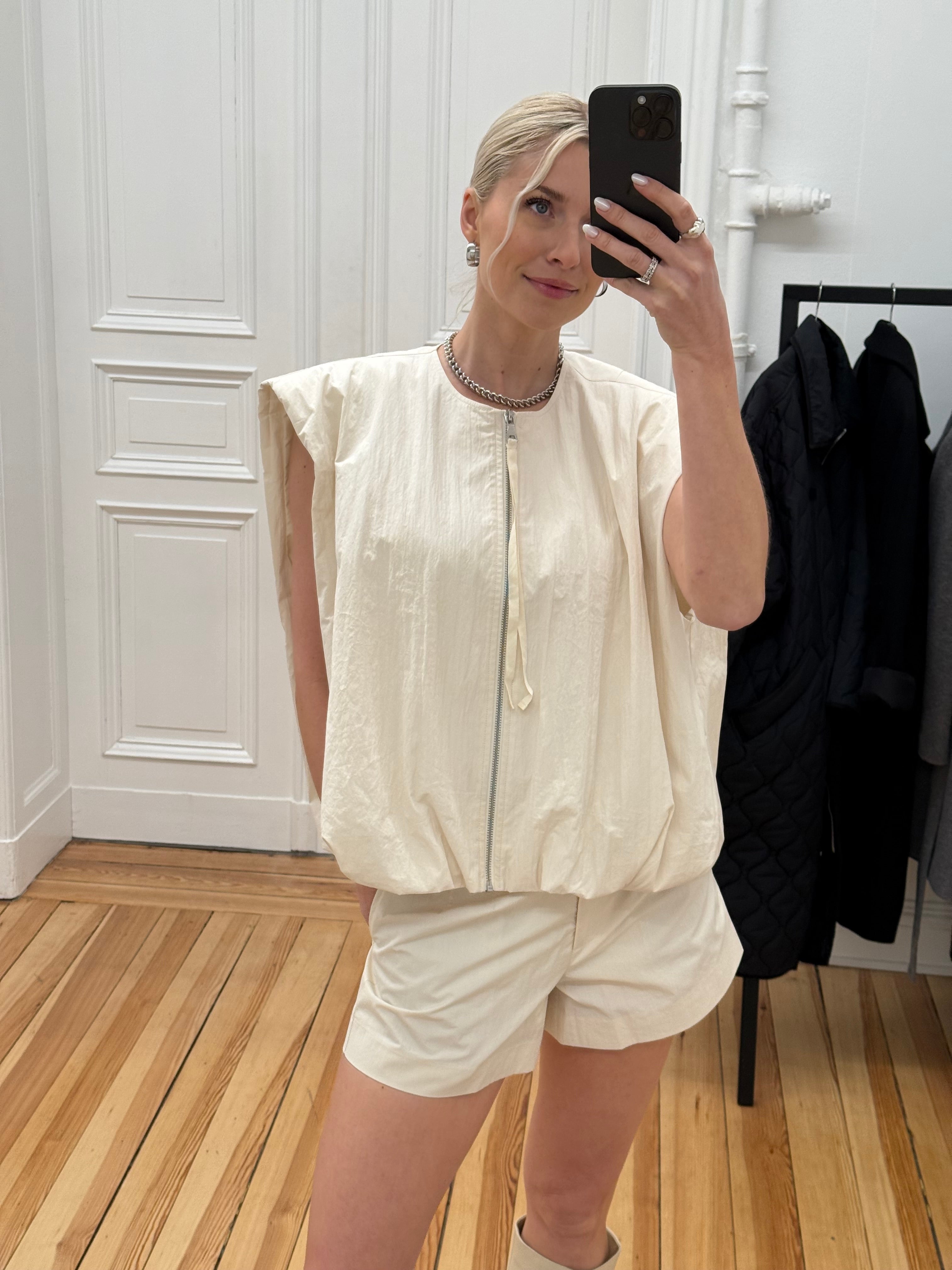 Eine Frau in der Marei Vest von LeGer by Lena Gercke steht auf einem Holzboden und macht ein Spiegel-Selfie in einem hellen, modernen Raum mit weißen Wänden und hängenden Mänteln im Hintergrund.