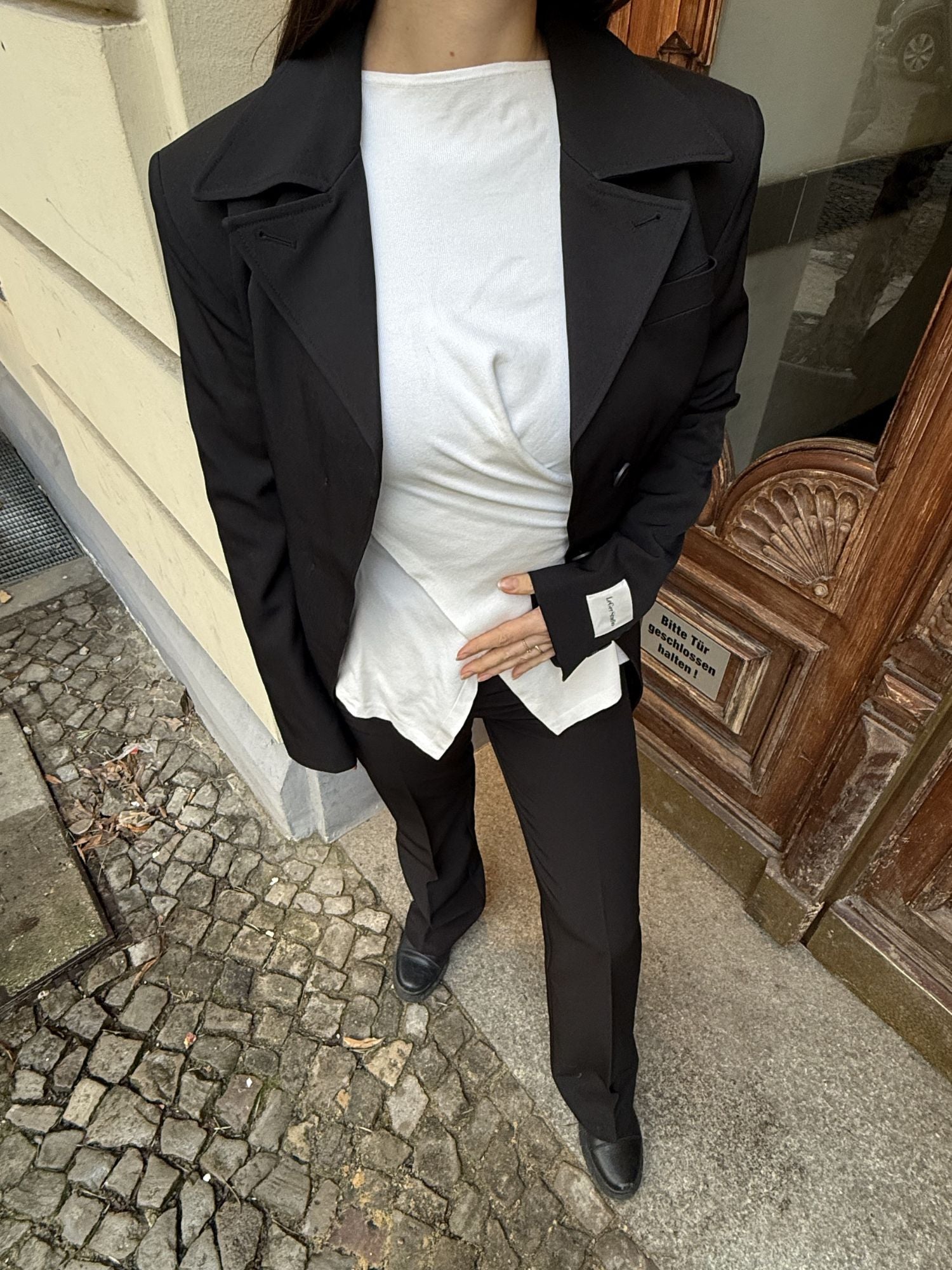 Eine Person in schwarzem Anzug und Schuhen über einem weißen Senta Longsleeve von LeGer by Lena Gercke steht auf einem kopfsteingepflasterten Bürgersteig in der Nähe eines cremefarbenen Gebäudes und rückt den Hemdsaum zurecht.