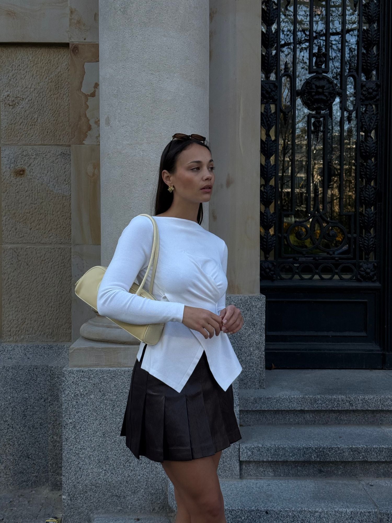 Eine Frau steht vor einem steinernen Gebäude mit einer verzierten Tür. Sie trägt das Senta Longsleeve von LeGer by Lena Gercke mit extra langen Ärmeln, einen braunen Faltenrock und eine hellgelbe Umhängetasche. Sie hat eine Sonnenbrille auf dem Kopf und blickt zur Seite.