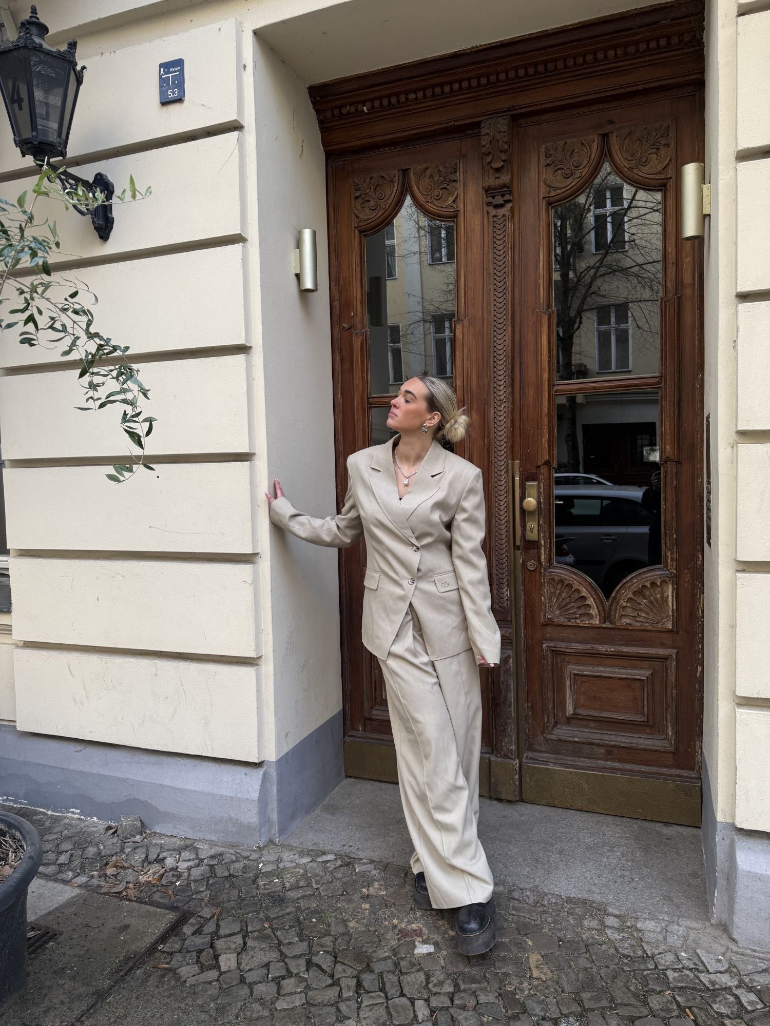 Eine Person in einem beigen Anzug mit weit geschnittenen Raquel Pants von LeGer by Lena Gercke und schwarzen Schuhen steht auf einem kopfsteingepflasterten Bürgersteig in der Nähe einer verschnörkelten Holztür neben einem cremefarbenen Gebäude.