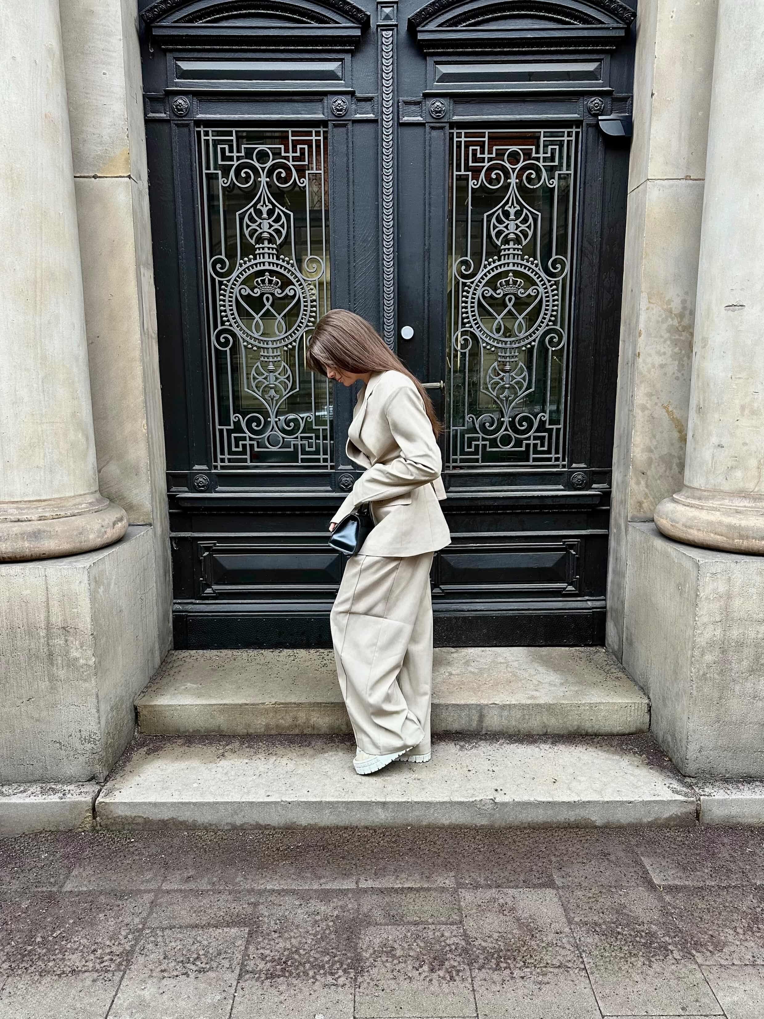 Eine Person, die die Raquel Pants von LeGer by Lena Gercke in Beige trägt, steht auf Steinstufen vor einer verzierten schwarzen Tür mit Eisenbeschlägen, hält eine schwarze Clutch in der Hand und blickt nach unten.
