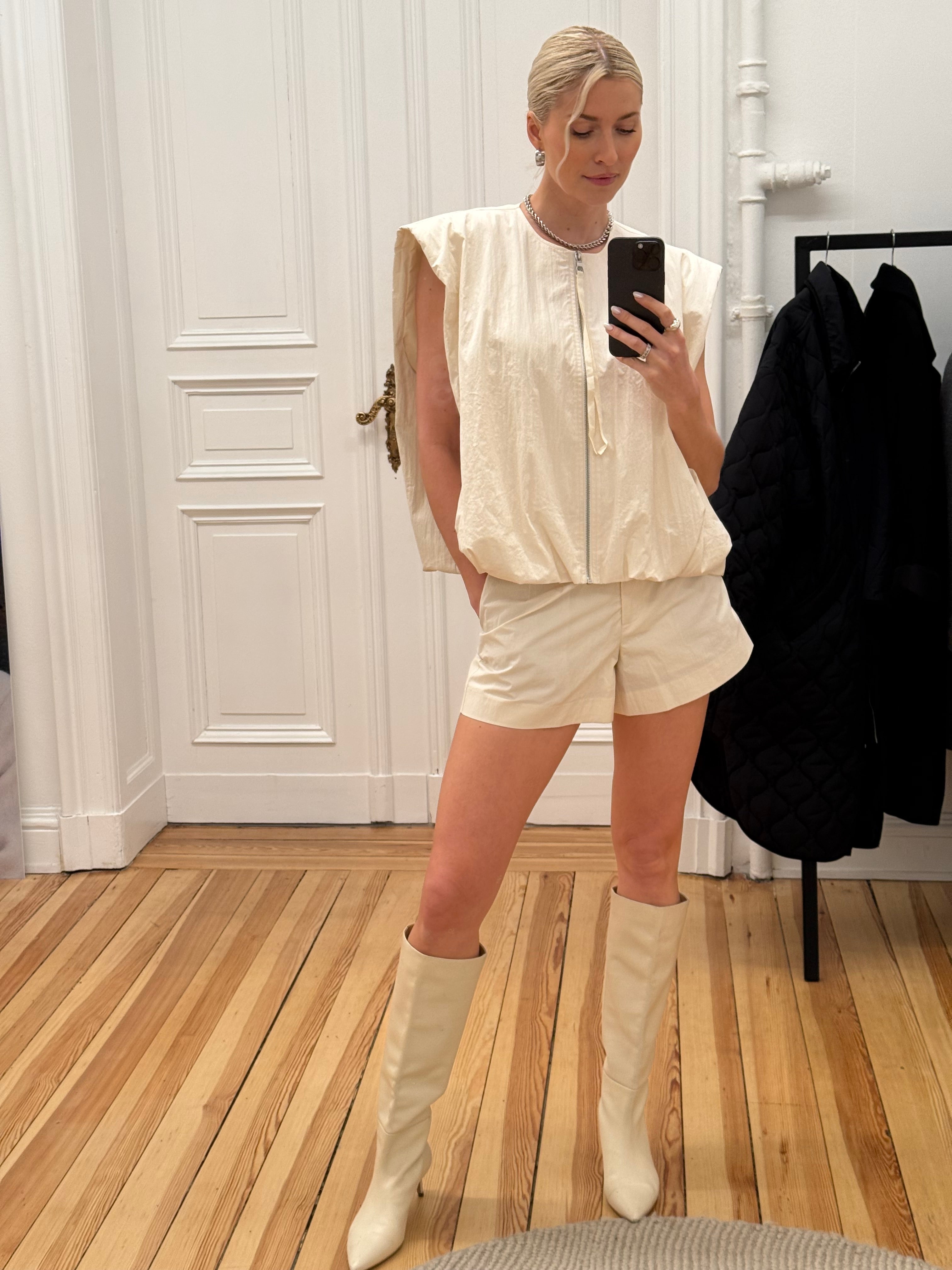 Eine Frau mit zurückgebundenen blonden Haaren macht ein Spiegel-Selfie in einem Innenraum. Sie trägt die Marei Shorts mit hoher Leibhöhe von LeGer by Lena Gercke, ein leichtes ärmelloses Top mit Reißverschluss und kniehohe cremefarbene Stiefel. Sie steht auf einem Holzfußboden neben einem schwarzen Mantel auf einem Ständer.
