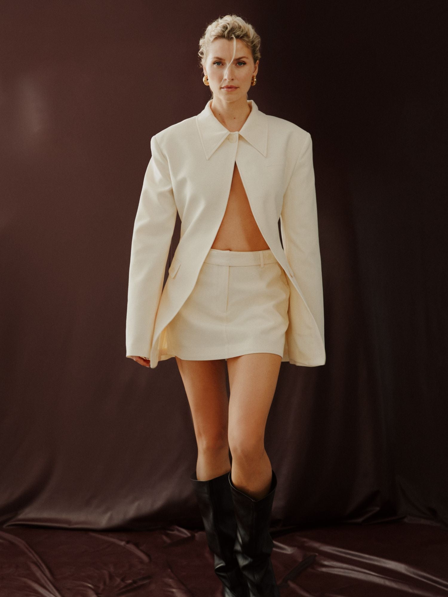 Eine Person posiert vor einem dunklen Hintergrund in einem cremefarbenen, nach vorne offenen Blazer und dem Cim Rock von LeGer by Lena Gercke, gestylt mit hohen schwarzen Stiefeln. Unter dem Blazer ist ihre Taille zu sehen. Sie hat kurzes, lockiges blondes Haar und einen selbstbewussten Blick.