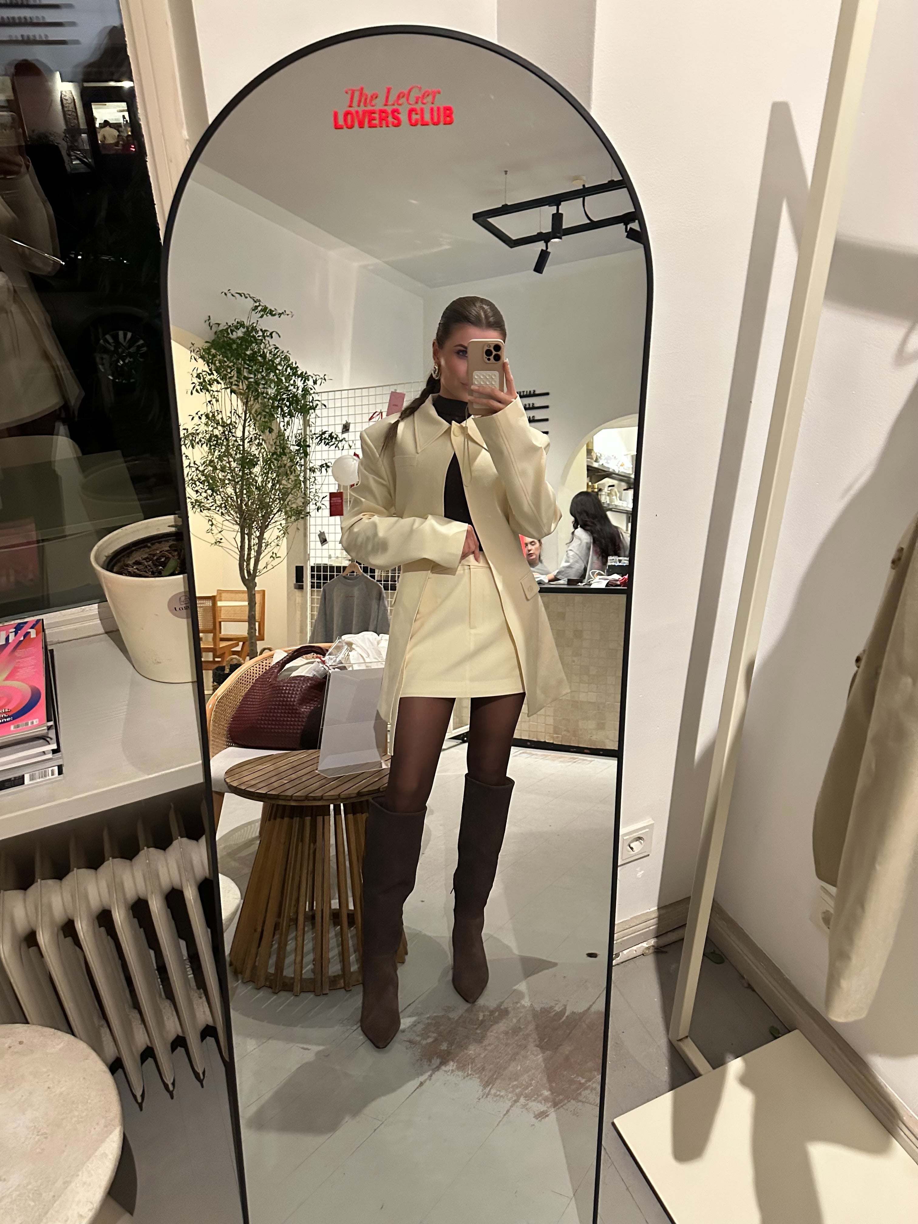 Eine Frau macht ein Spiegel-Selfie in einem stilvollen Zimmer und trägt einen cremefarbenen LeGer by Lena Gercke Cim Rock, einen passenden Mantel, ein schwarzes Top und hohe braune Stiefel. Oben auf dem Spiegel steht in roter Schrift "The LOFT LOVERS CLUB".