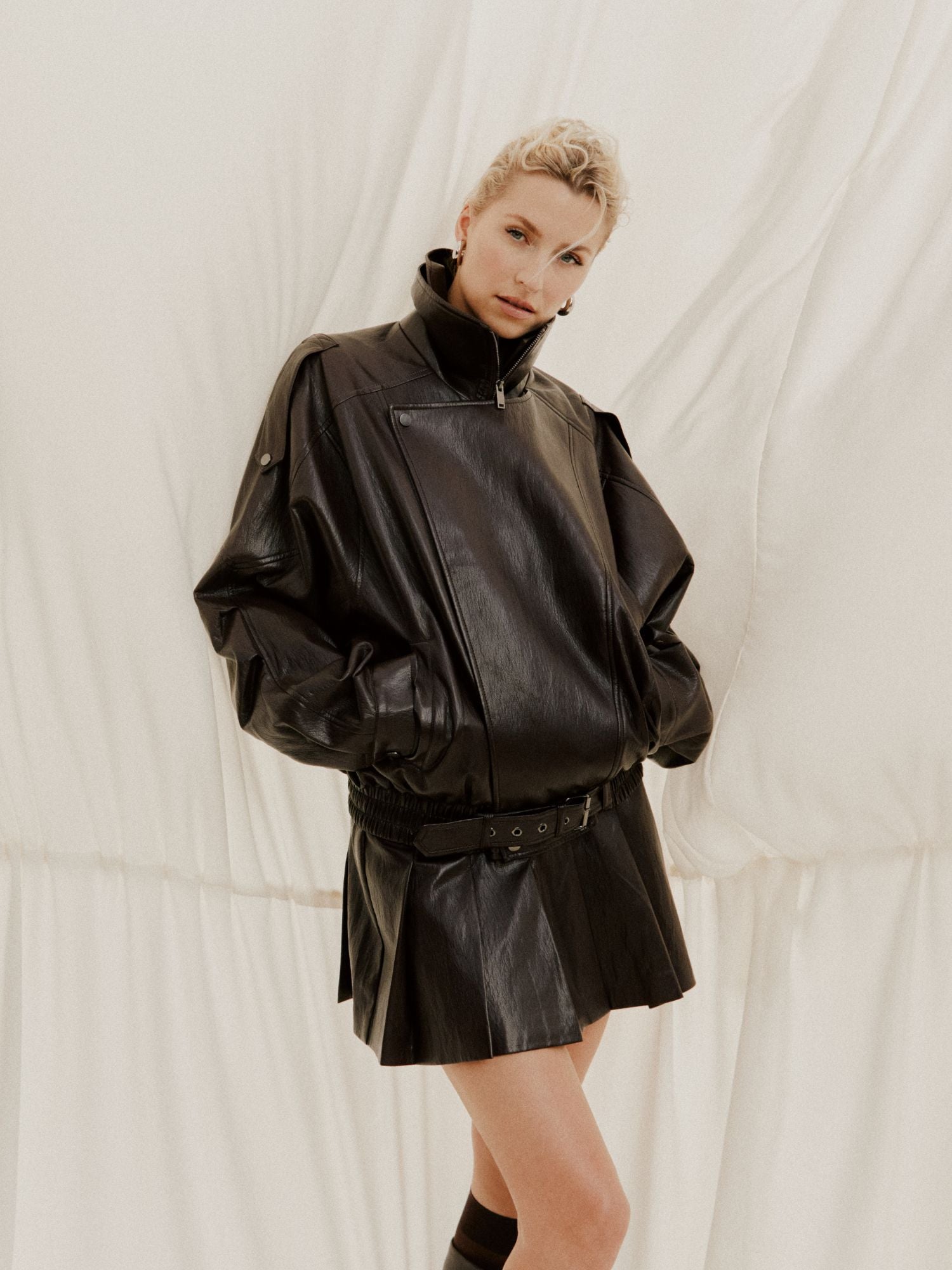 Vor einem cremefarbenen Hintergrund steht eine Person, die selbstbewusst eine übergroße Jacke von Leder-Optik und den Jessa Rock von LeGer by Lena Gercke trägt, die Hände in den Taschen und den Blick keck in die Kamera gerichtet.