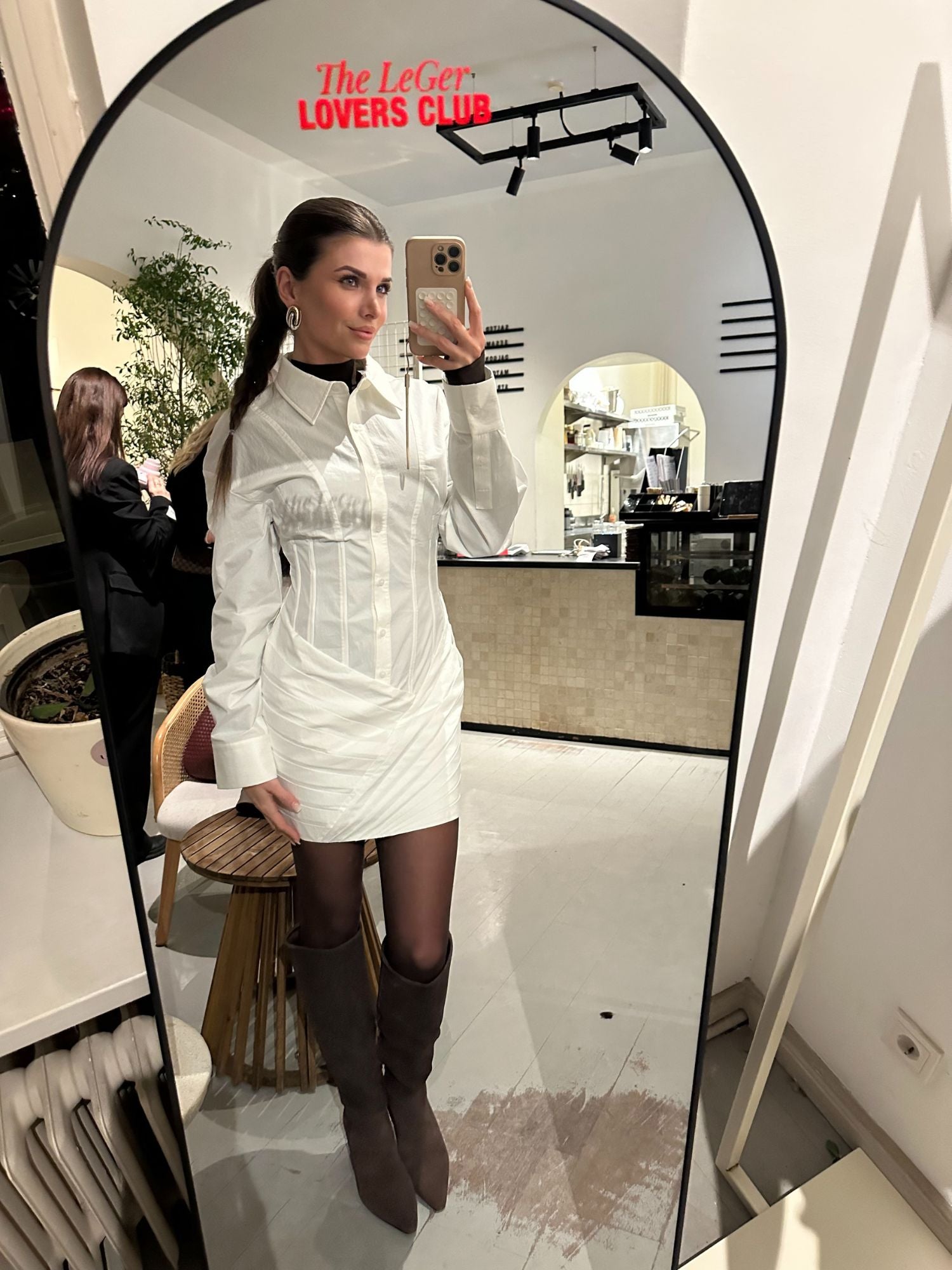 Eine Frau in einem weißen Esma-Kleid von LeGer by Lena Gercke, gestylt mit dunkler Strumpfhose und kniehohen braunen Stiefeln, knipst ein Spiegel-Selfie in einem schicken Raum mit Pflanzen. Oben auf dem Spiegel steht in roter Schrift "The LeGer LOVERS CLUB".